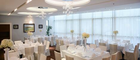 A banquet hall with round tables, white tablecloths, and chairs, featuring a chandelier and large windows.
