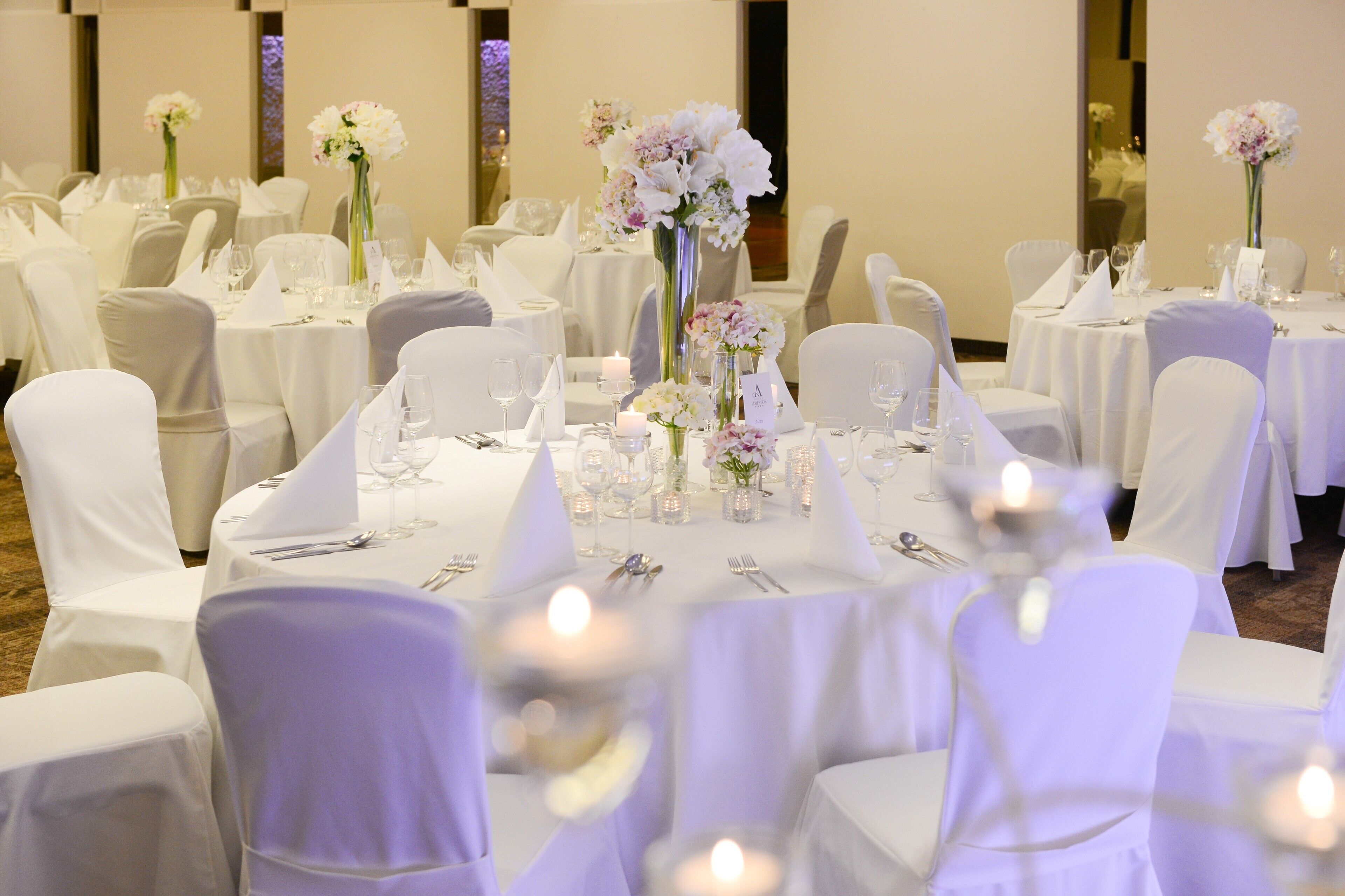 Une salle de banquet avec des tables rondes dressées pour le repas, des nappes blanches et des chaises recouvertes d’un tissu blanc.