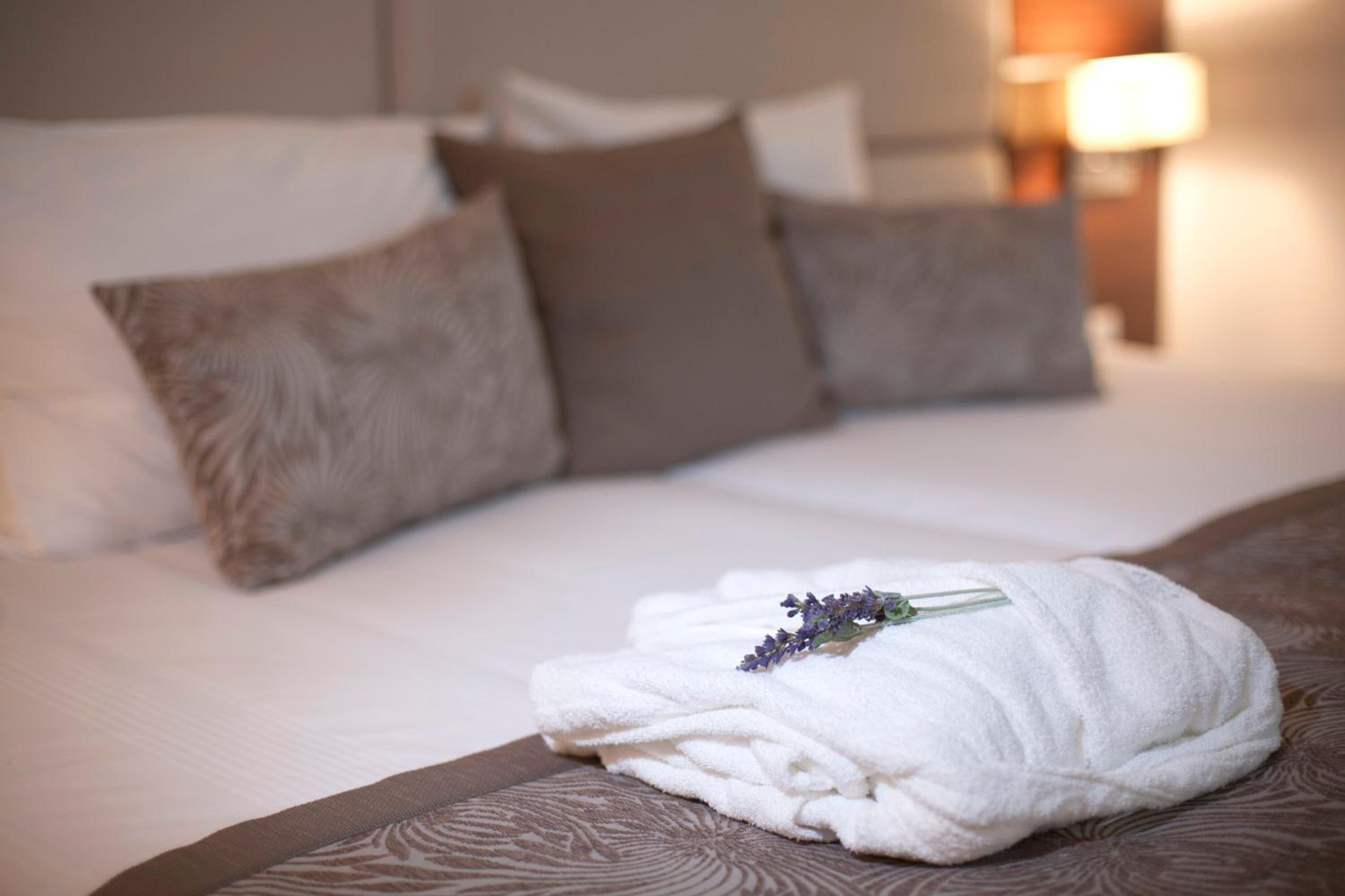 A neatly made bed with a white towel and a lavender flower, surrounded by brown pillows and a patterned bedspread.