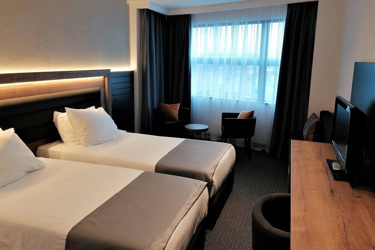 A hotel room with two beds, a desk, a chair, a TV, and a window with curtains.