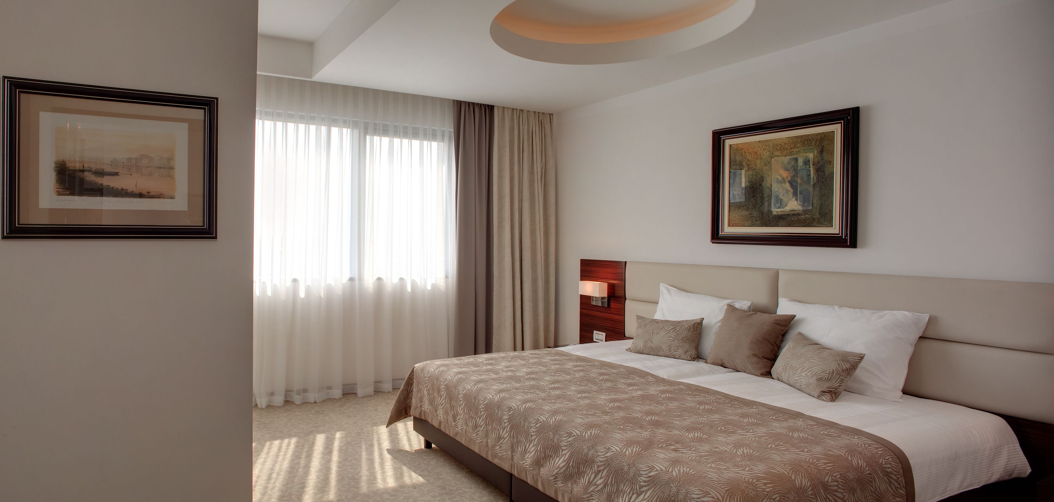 A hotel room with a large bed, a headboard with a light fixture, and two framed artworks on the walls.