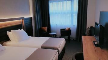 Een hotelkamer met twee bedden, een kleine tafel, een stoel, een televisie en uitzicht op het omliggende landschap door dunne gordijnen.
