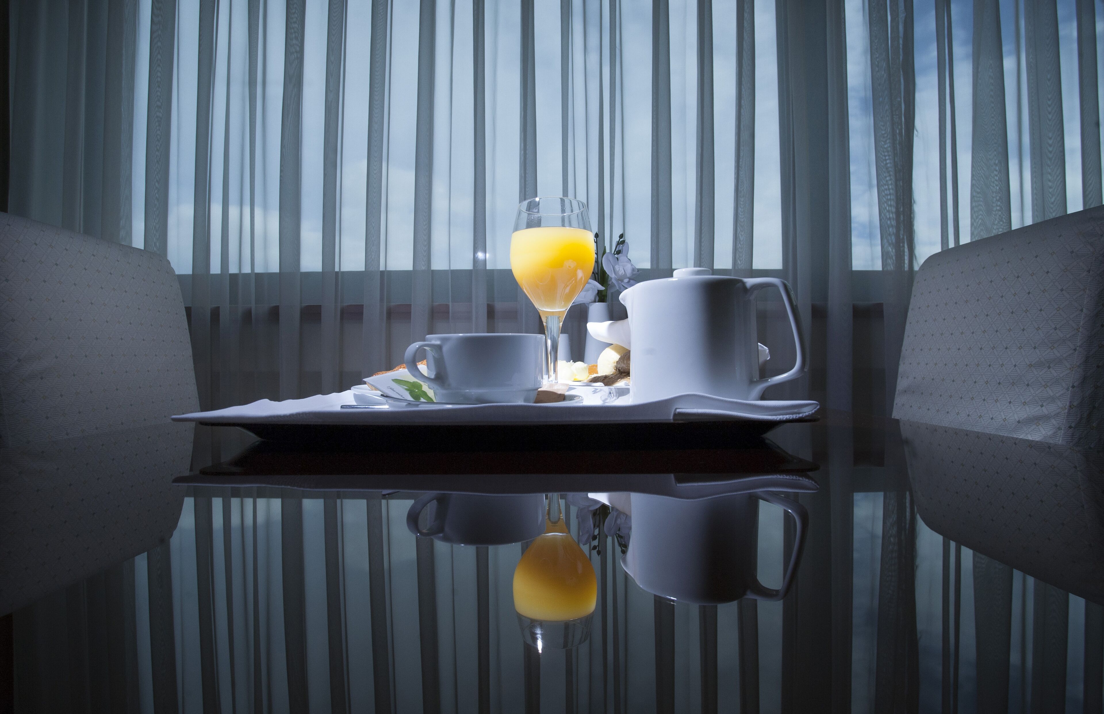 A breakfast tray with a glass of orange juice, a cup, and a pitcher on a reflective table in front of a window with sheer curtains.