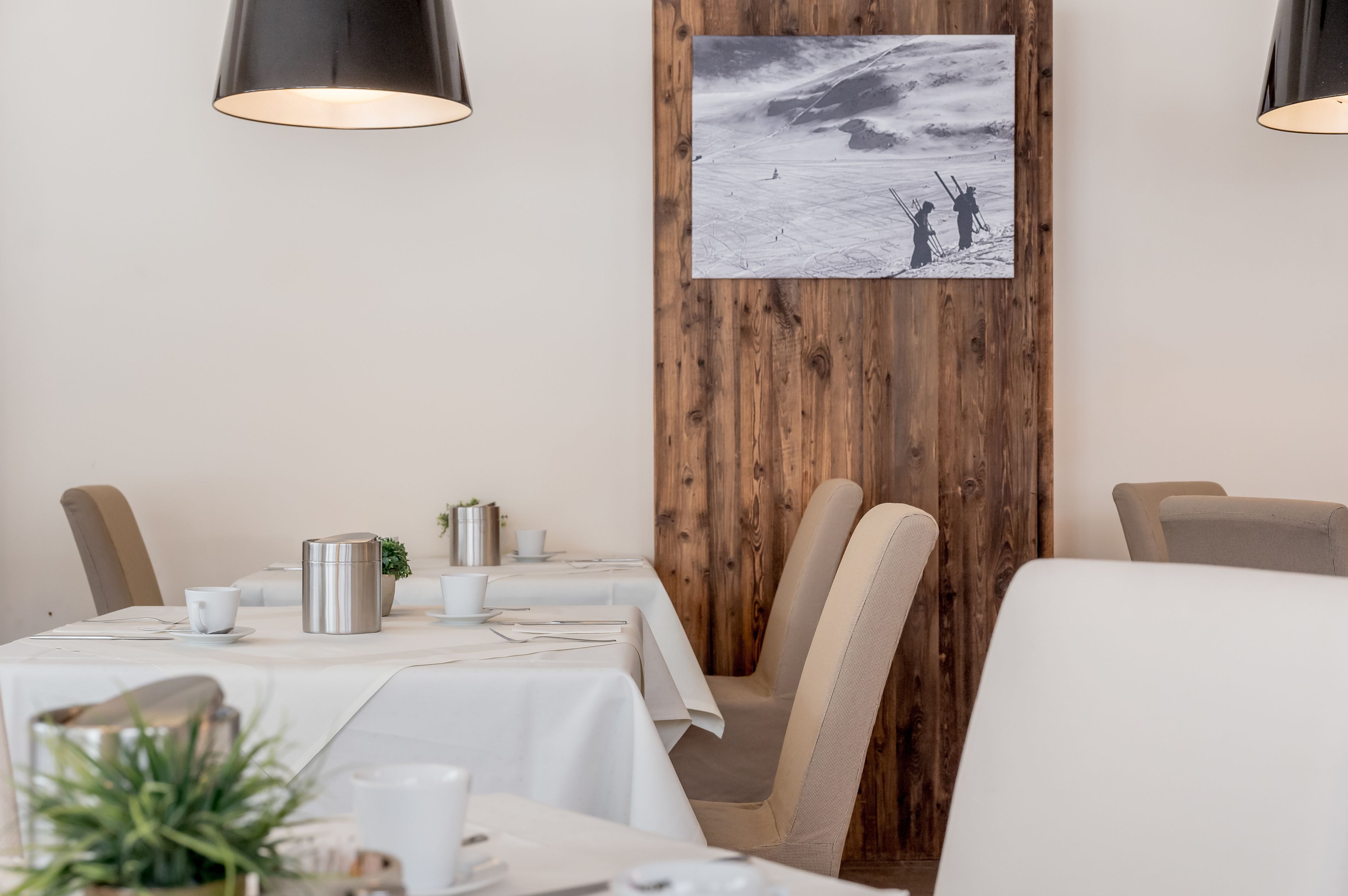 Un comedor con mesas dispuestas para dos, cada una con mantel blanco, cubiertos y una planta pequeña. Dos lámparas colgantes se encuentran suspendidas sobre las mesas.