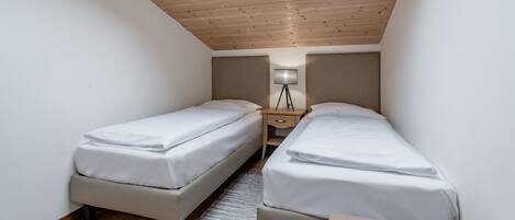 Una habitación pequeña y luminosa con dos camas, una mesita de noche, una lámpara y un tragaluz.
