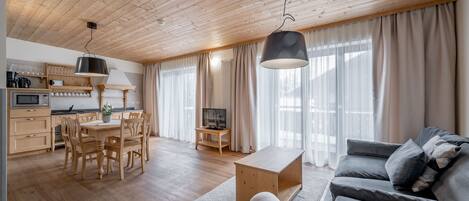 A modern living space with a wooden ceiling, a dining area with a wooden table and chairs, a grey sofa, and a television on a stand.
