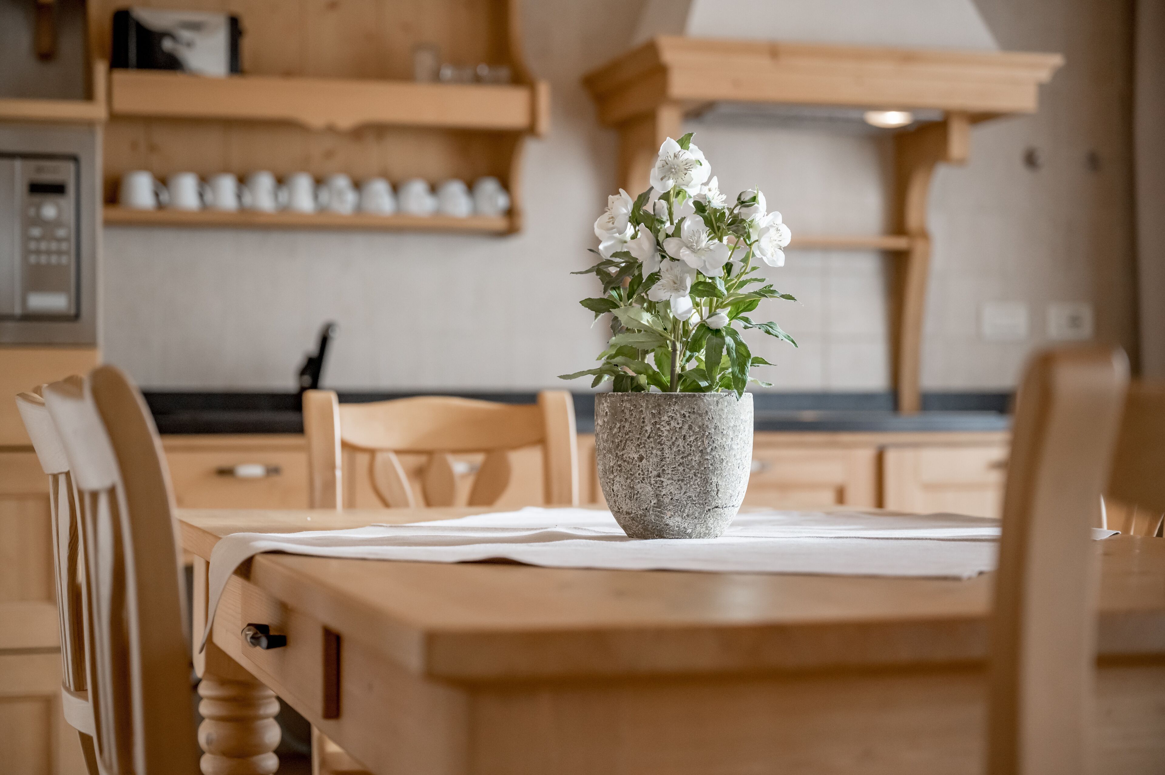 Una mesa de comedor de madera con un florero de piedra que contiene flores blancas, ubicada en una cocina con armarios de madera y una máquina de café al fondo.