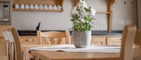 A wooden dining table with a stone vase containing white flowers, set in a kitchen with wooden cabinets and a coffee machine in the background.