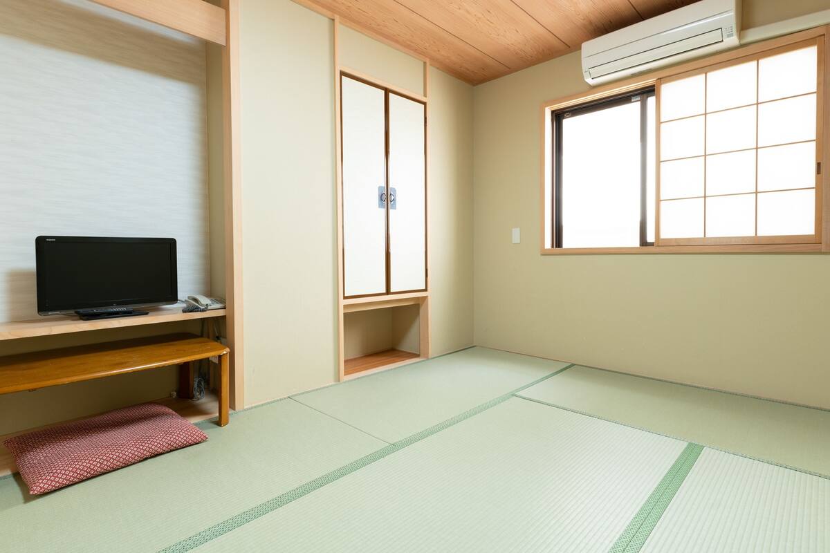 Een kamer met tatami vloerbedekking, een televisie op een houten standaard, een schuifdeur en een raam met een rasterpatroon.