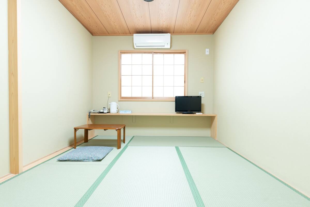 Een kamer met tatami vloerbedekking, een lage houten tafel, een televisie op een plank en een raam met rasterwerk.