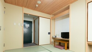 Een traditionele Japanse kamer met tatami vloerbedekking, een schuifdeur, een televisie op een houten standaard en een klein tafeltje met een kussen.