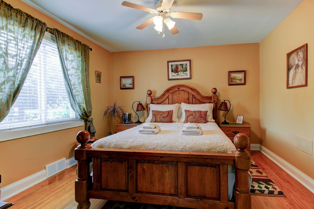 A bedroom with a wooden bed, a ceiling fan, a window with curtains, and framed pictures on the walls.
