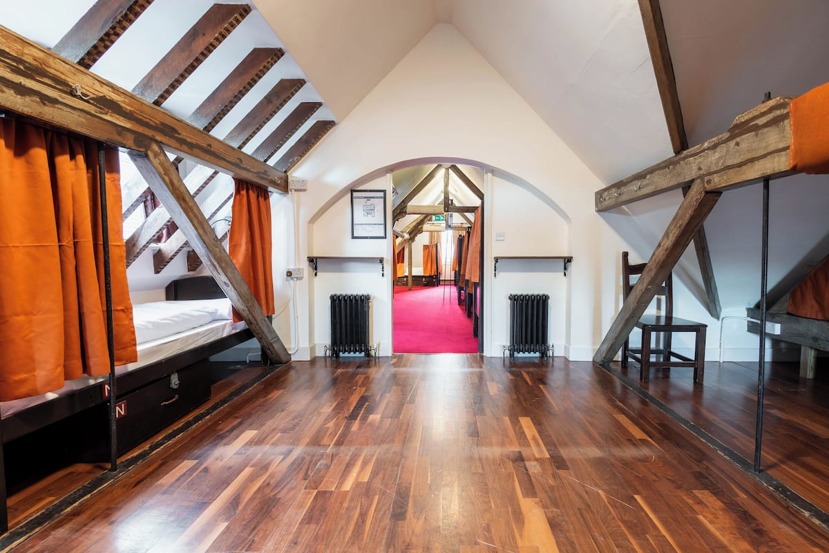 A loft bedroom with wooden beams, a red carpet, and a bed with orange curtains.