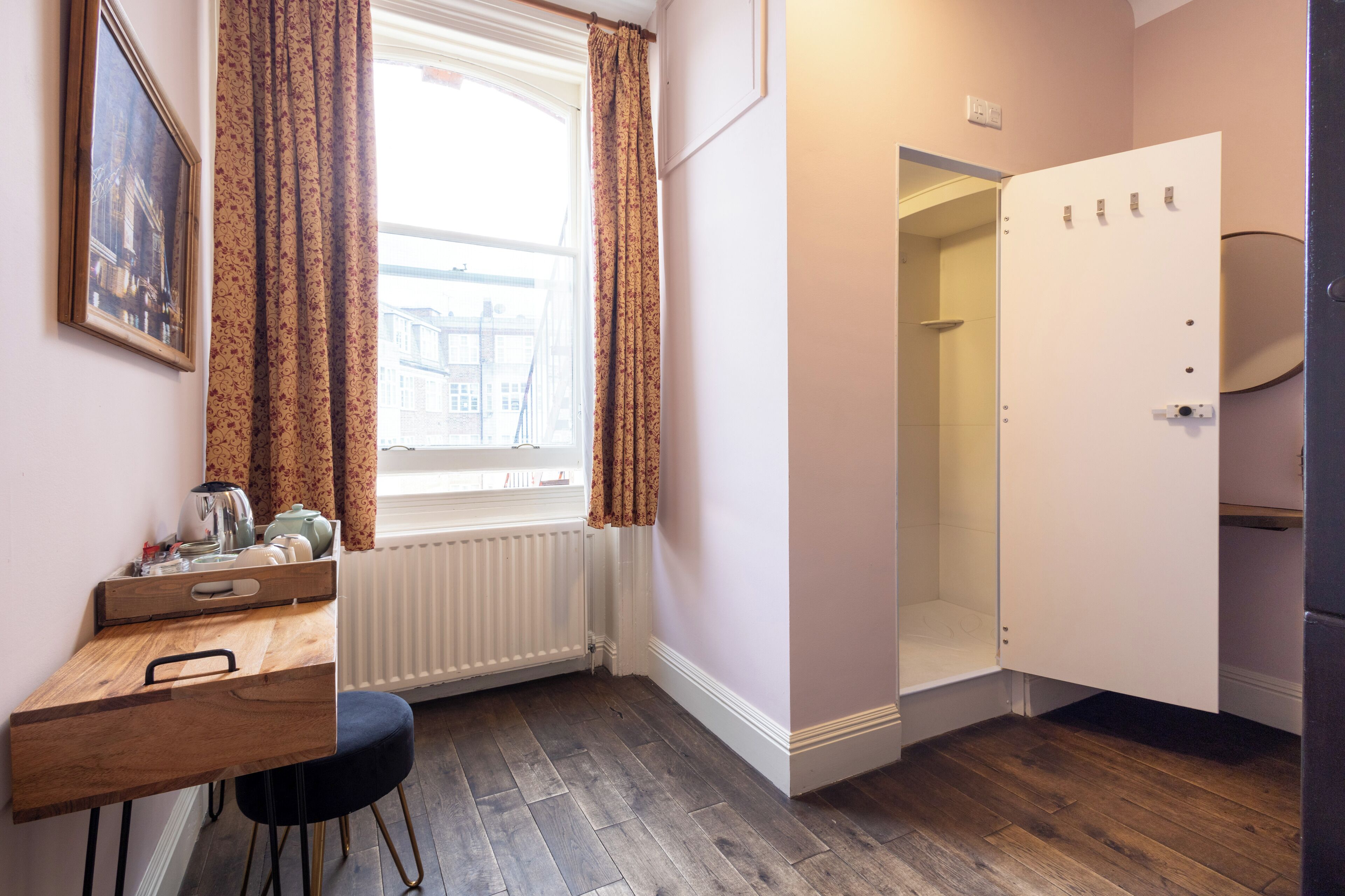 A compact room with a wooden desk, a radiator, a window with curtains, and a white door leading to a bathroom.
