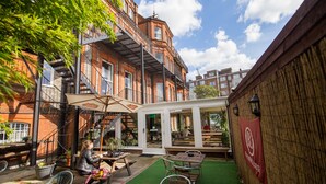 Un patio al aire libre con mesas y sillas, una zona de estar cubierta y vistas a un edificio de ladrillo con varias ventanas y una escalera.