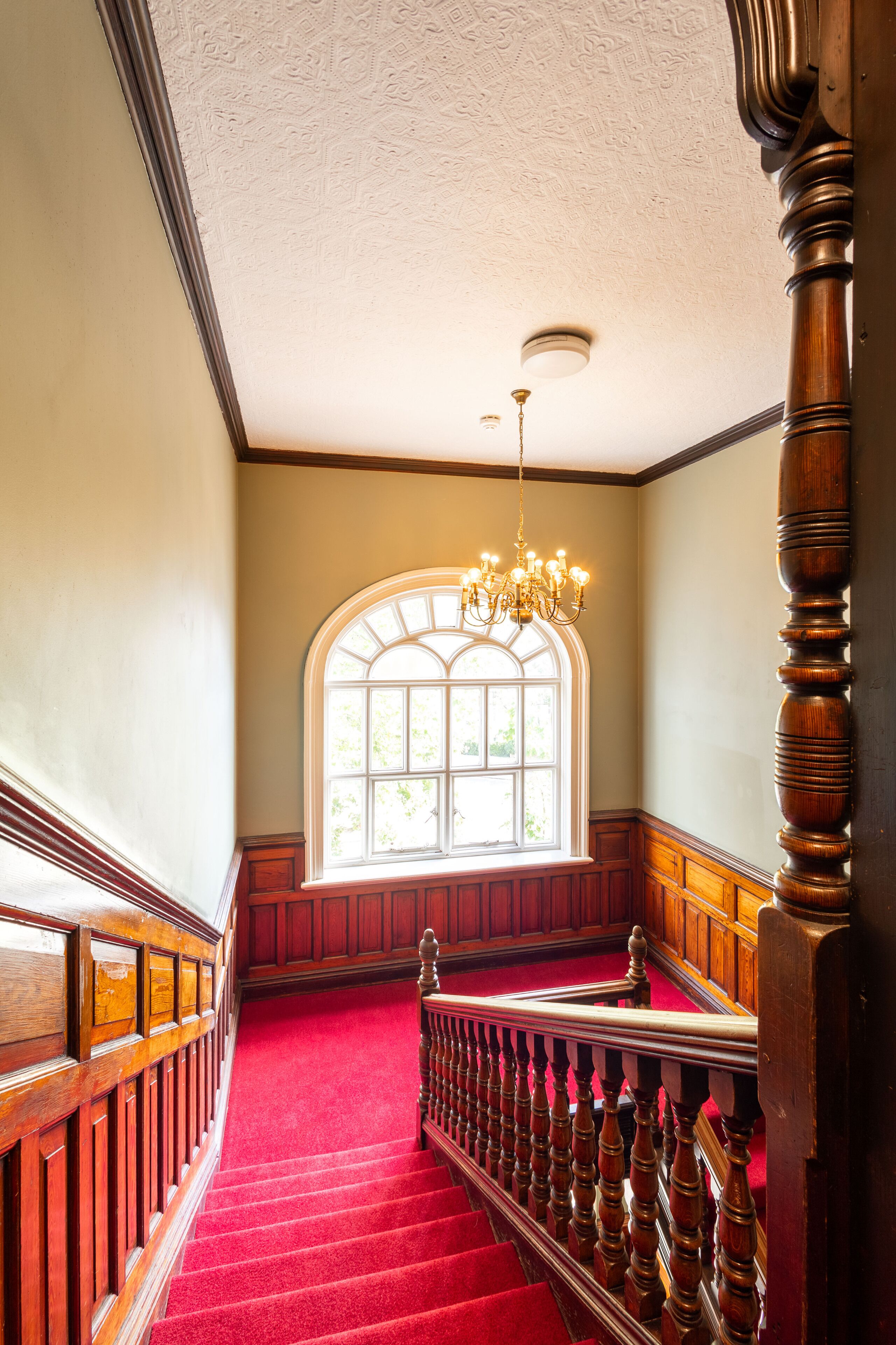 A grand staircase with a red carpet, wooden balustrades, and a large arched window with natural light.