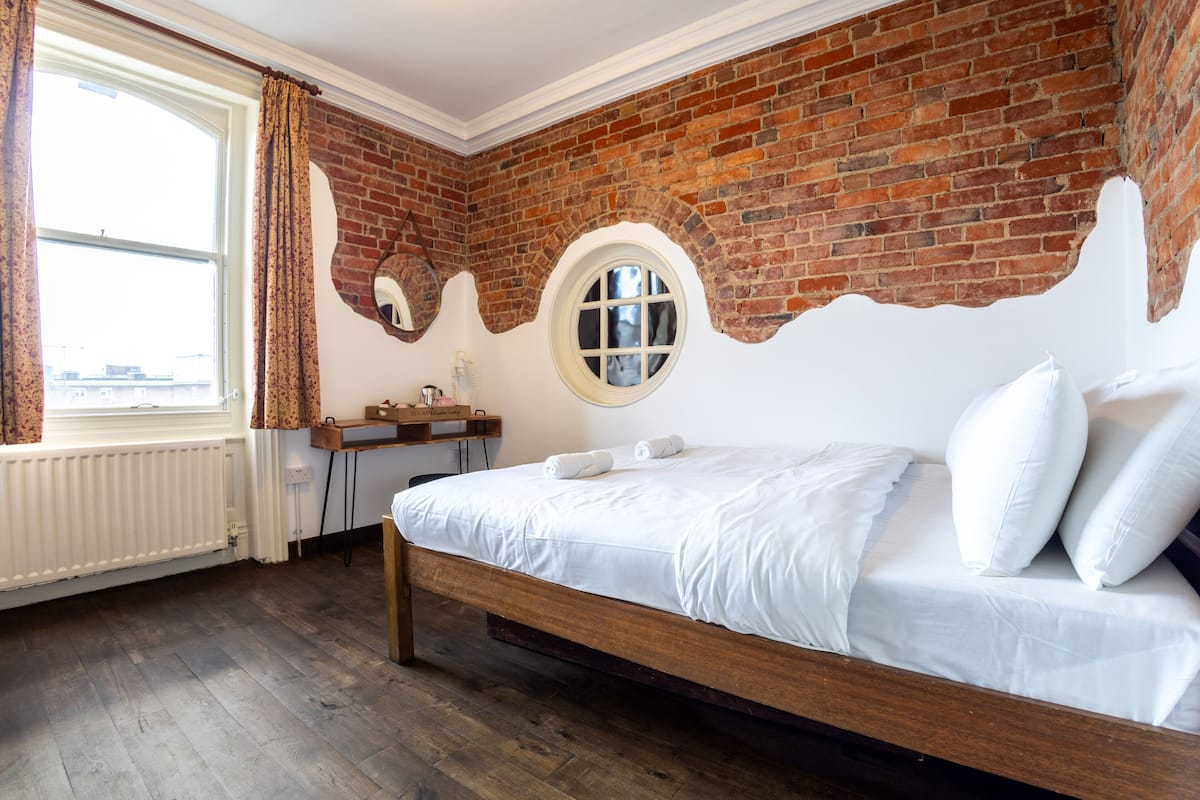 A bedroom with a brick wall, a wooden bed, a round window, and a radiator.