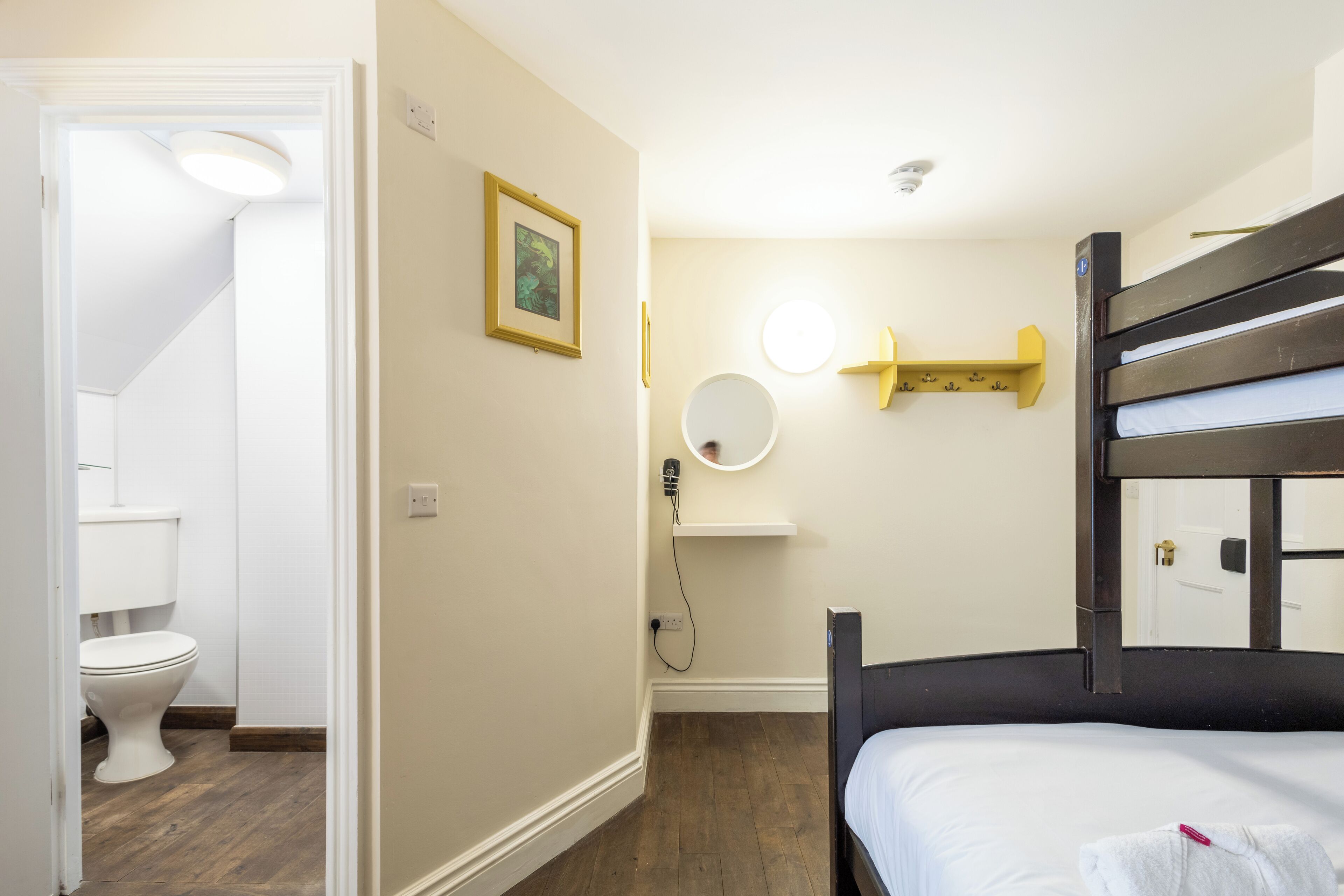 A hotel room with a bunk bed, a small bedside table, a mirror, and a shelf with toiletries.