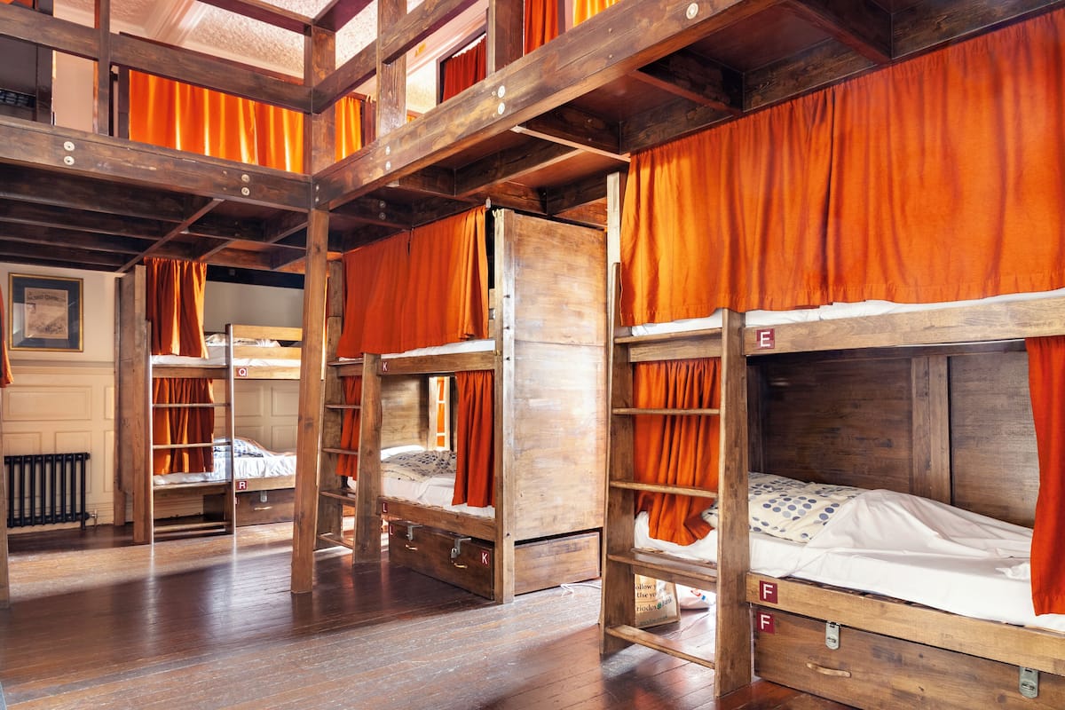 A dormitory-style room with bunk beds, wooden beams, and orange curtains.