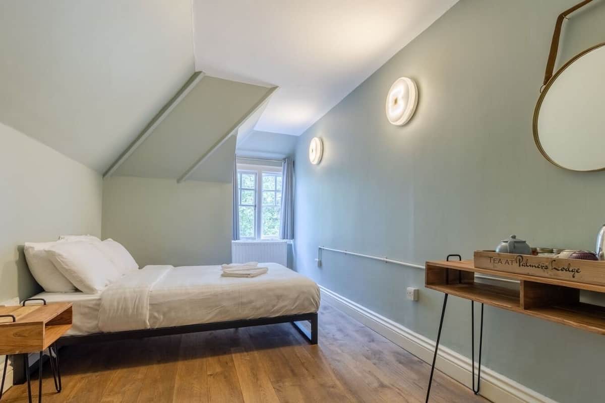 A bedroom with a bed, a wooden side table, a wall-mounted mirror, and a shelf with tea and kitchen items.