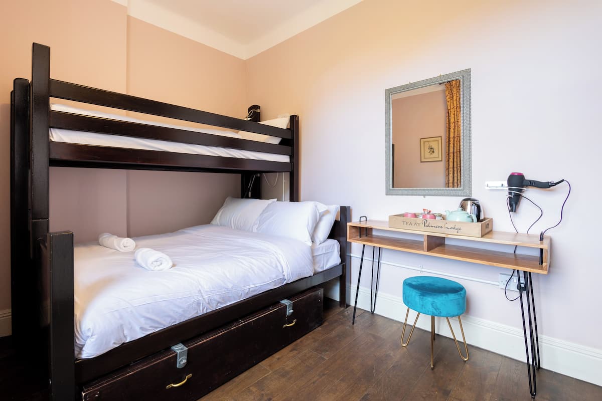 A bunk bed with white bedding, a wooden desk with a mirror, and a blue stool.