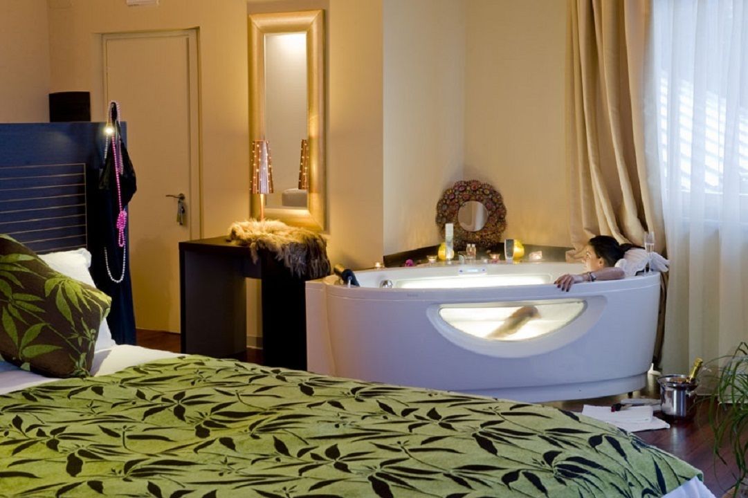 Una habitación de hotel con cama, un espejo grande, una bañera y una ventana con cortinas.