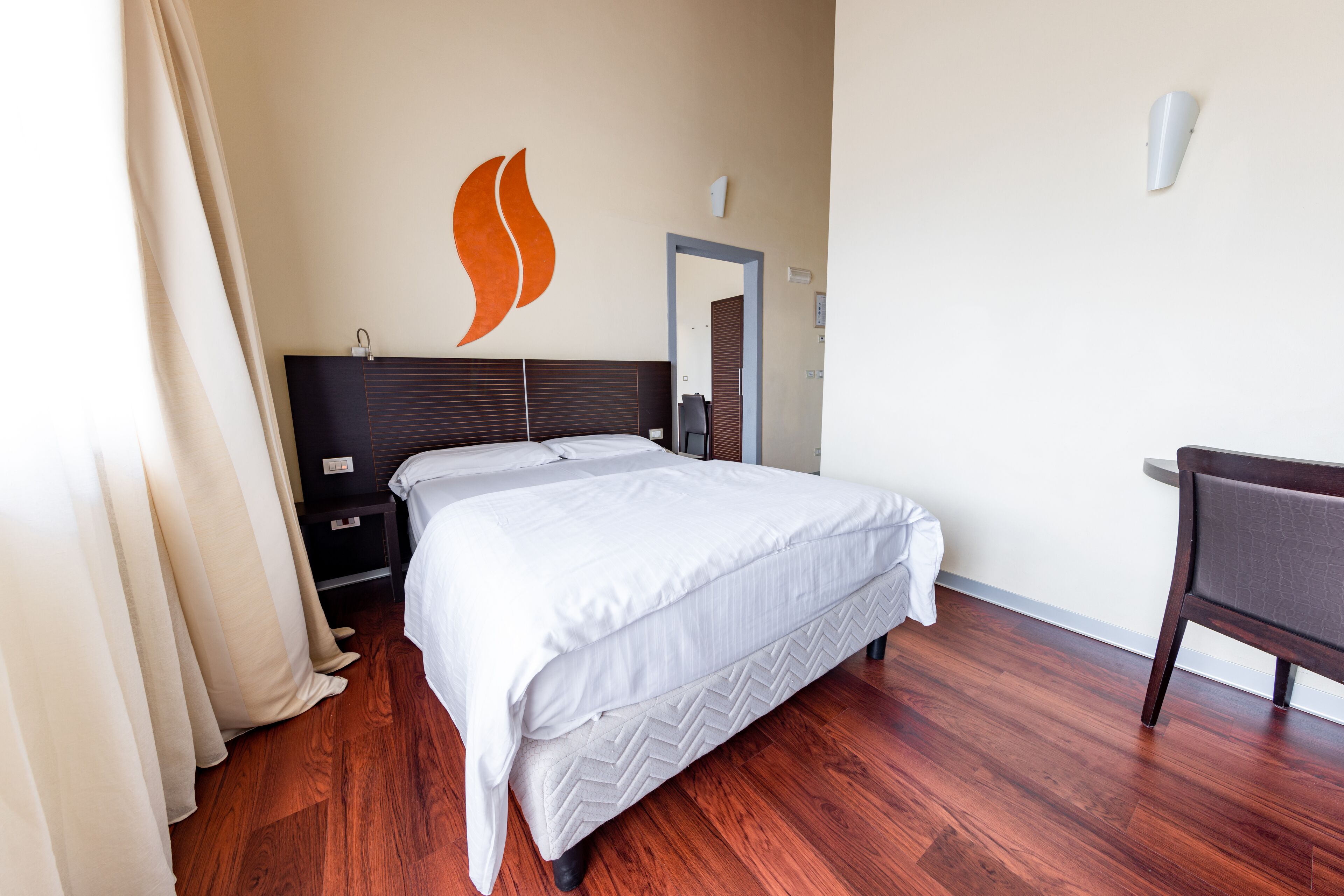 Habitación de hotel ordenada con una cama grande, una silla, una mesita de noche y un aplique de pared.