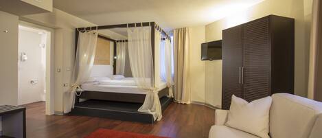 A bedroom with a four-poster bed, a sofa, a TV mounted on the wall, and a wardrobe.