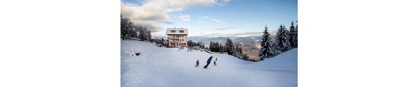 Una ladera cubierta de nieve con un edificio al fondo, rodeada de árboles cargados de nieve.