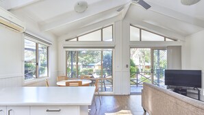A spacious room with a high ceiling, large windows, a ceiling fan, a flat-screen TV, and a dining area with chairs and a table. - NRMA Ocean Beach Holiday Resort (Umina Beach)