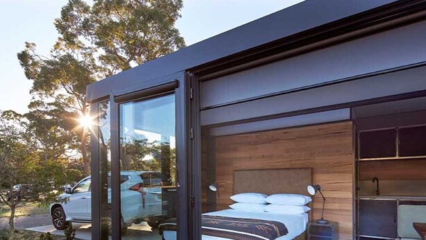 A modern bedroom with a large bed, wooden headboard, and a glass door leading to a driveway. - NRMA Ocean Beach Holiday Resort (Umina Beach)