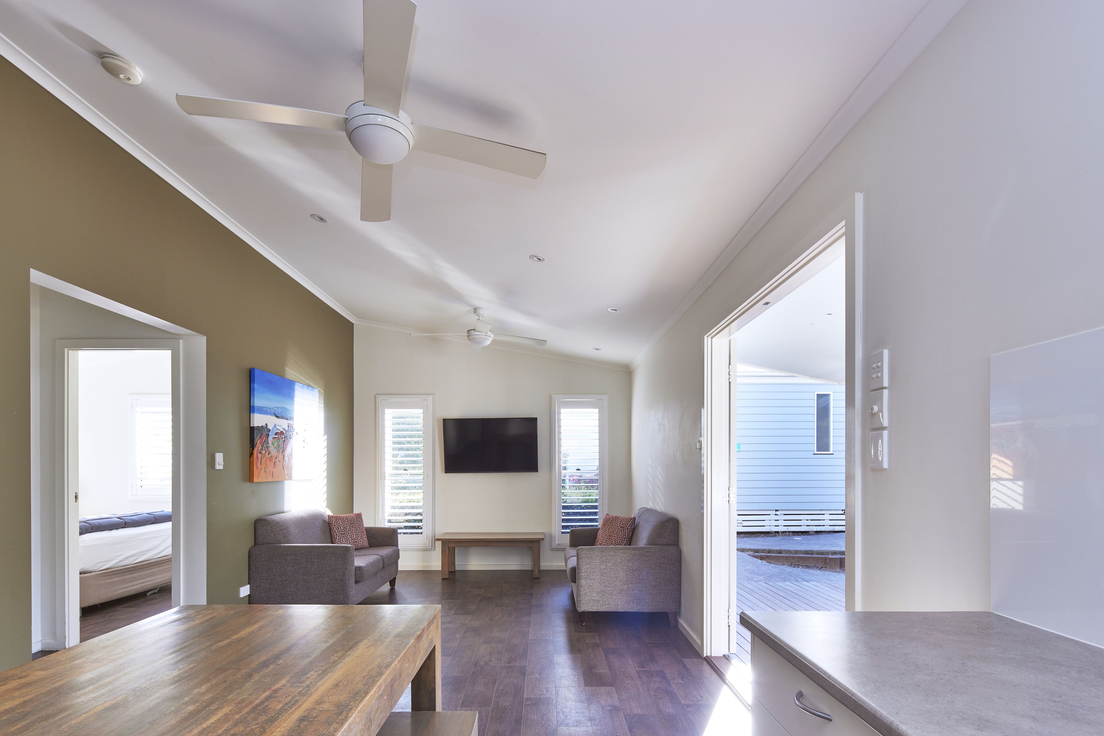 A spacious living room with a ceiling fan, a flat-screen TV mounted on the wall, and a large window providing natural light.