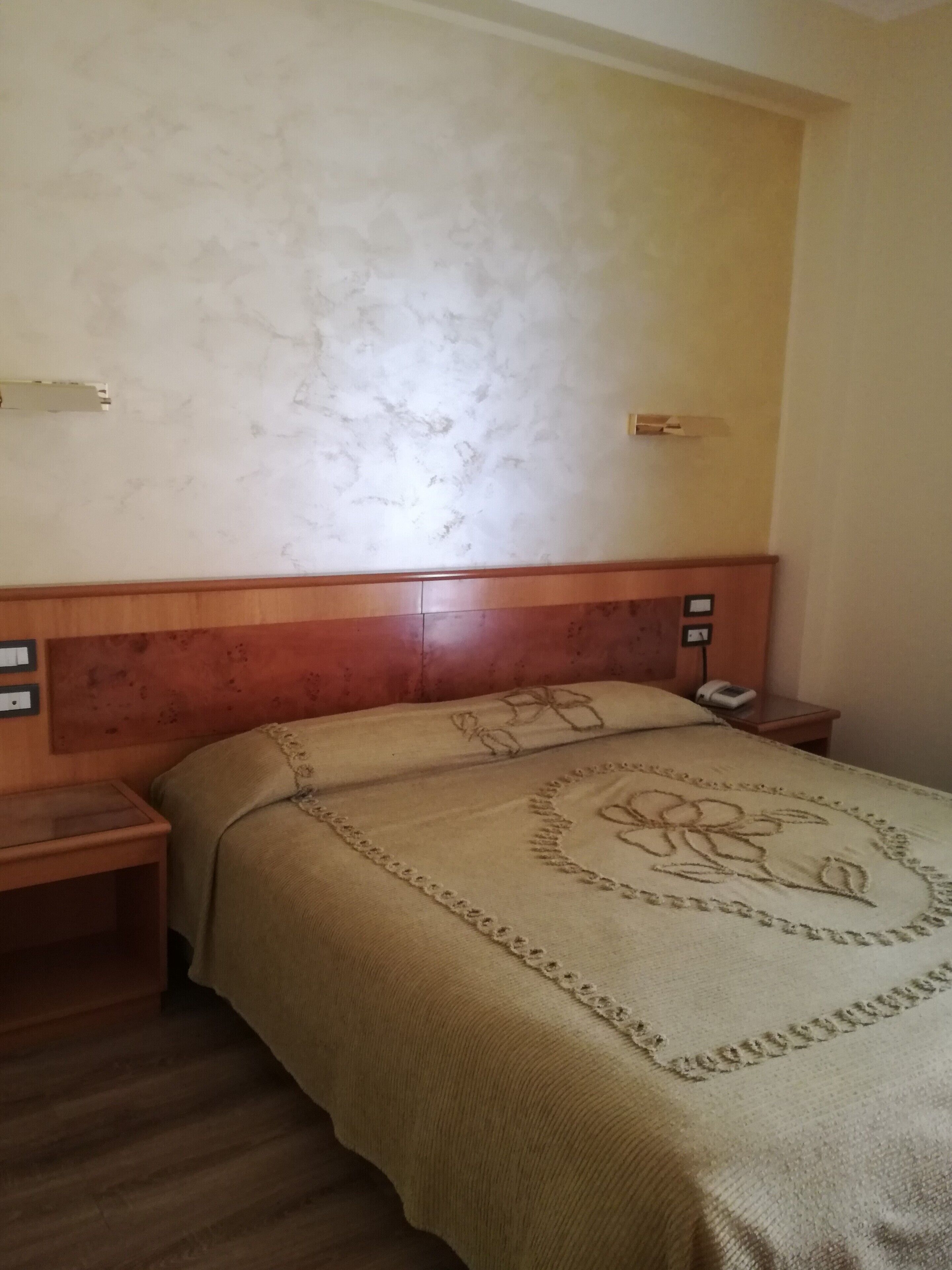Quarto de hotel com cabeceira de madeira, cama com colcha floral, mesa de cabeceira com telefone e interruptor de luz na parede.
