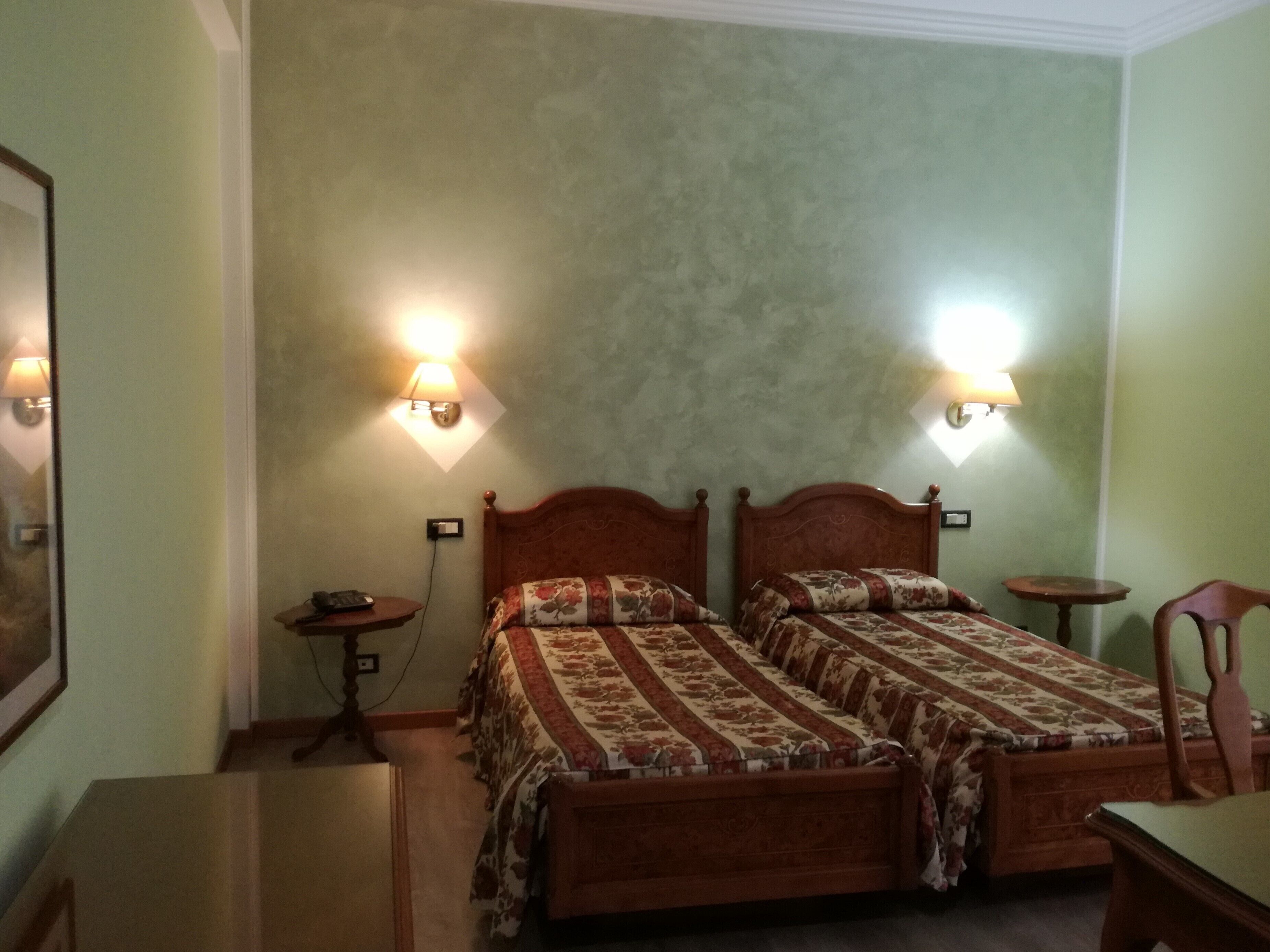 A room with two wooden beds, each with floral bedding, two bedside lamps, a small wooden table, and a chair.