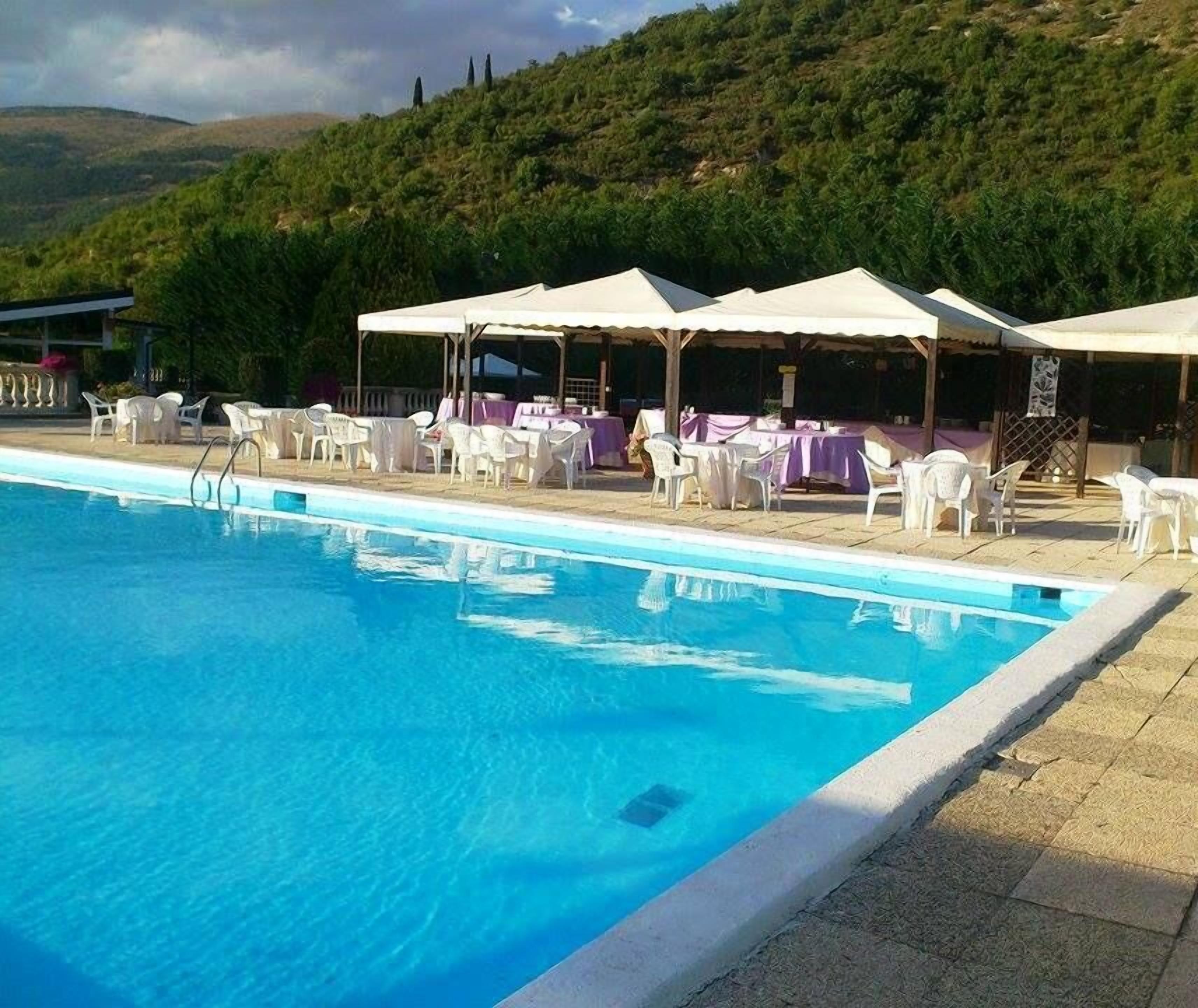 A swimming pool with white lounge chairs and tables, surrounded by a covered outdoor area with umbrellas.
