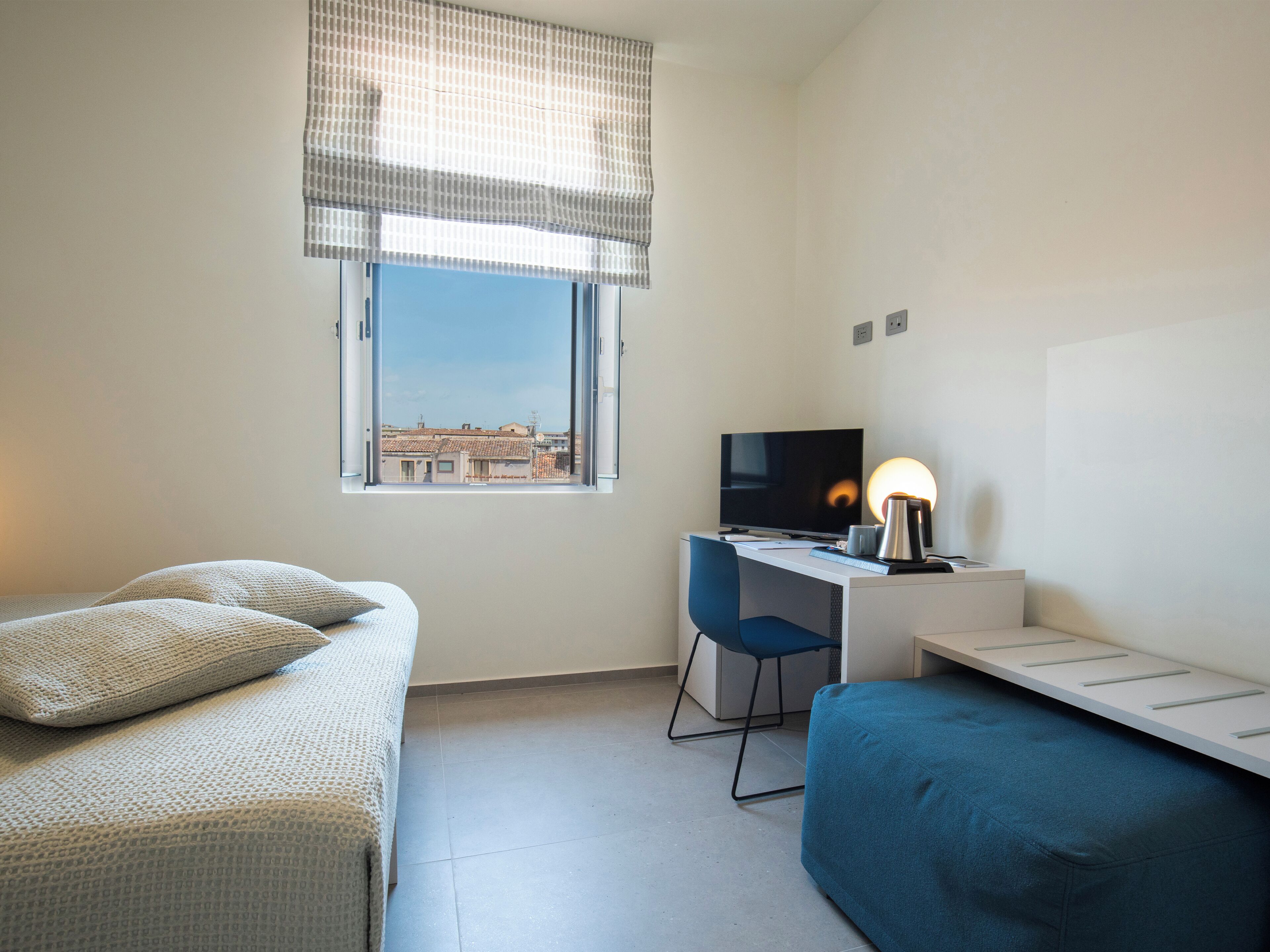 A bedroom with a bed, a desk with a computer, a chair, and a window with a view of buildings.