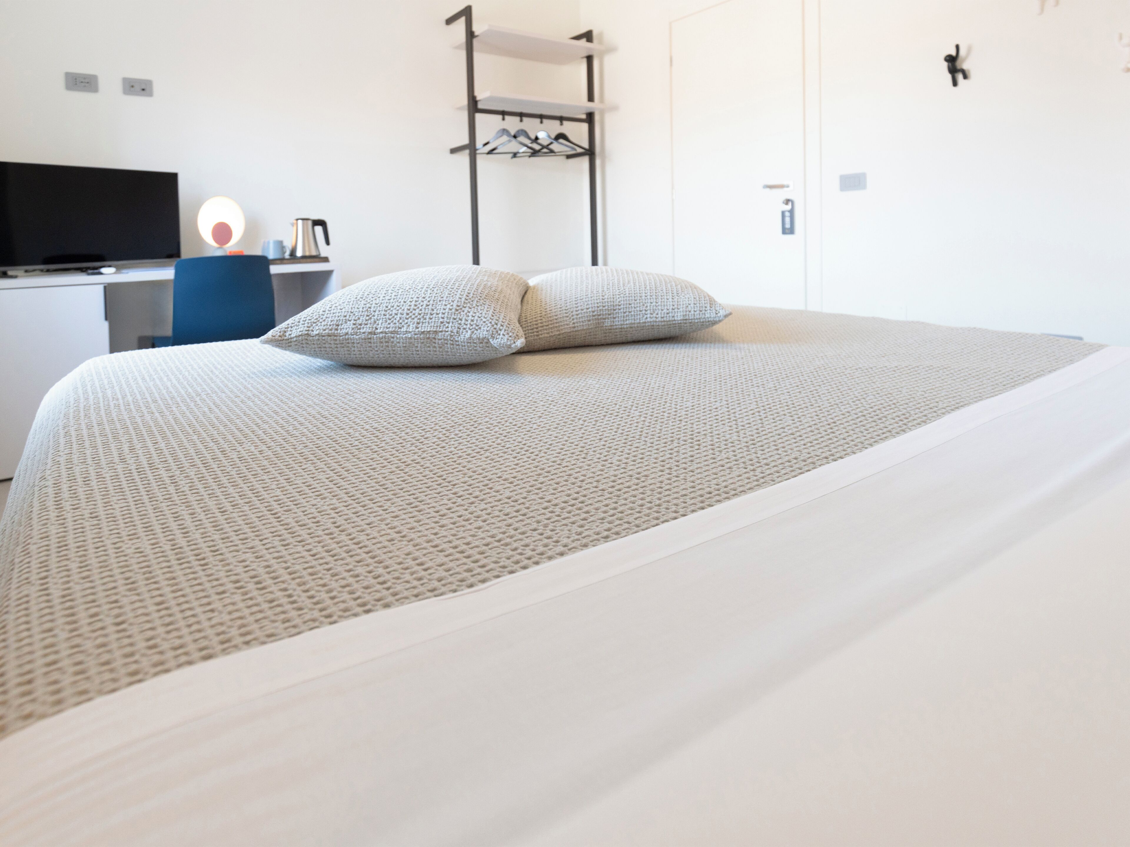 A neatly made bed with a textured white bedspread and two pillows, a desk with a computer and a lamp, and a wall-mounted coat rack.