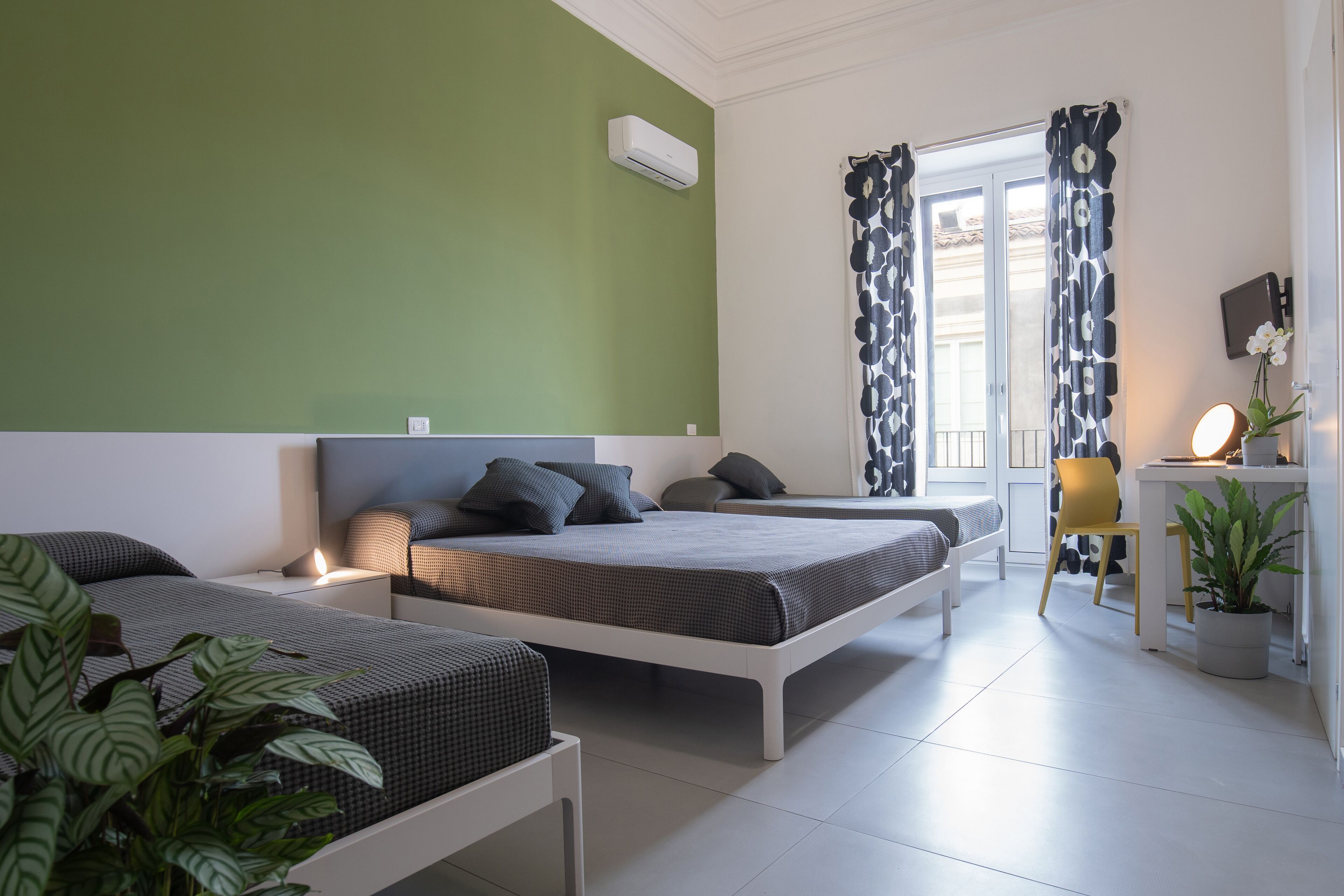 Un dormitorio con dos camas, un escritorio con silla, una planta en maceta y una ventana con cortinas estampadas.