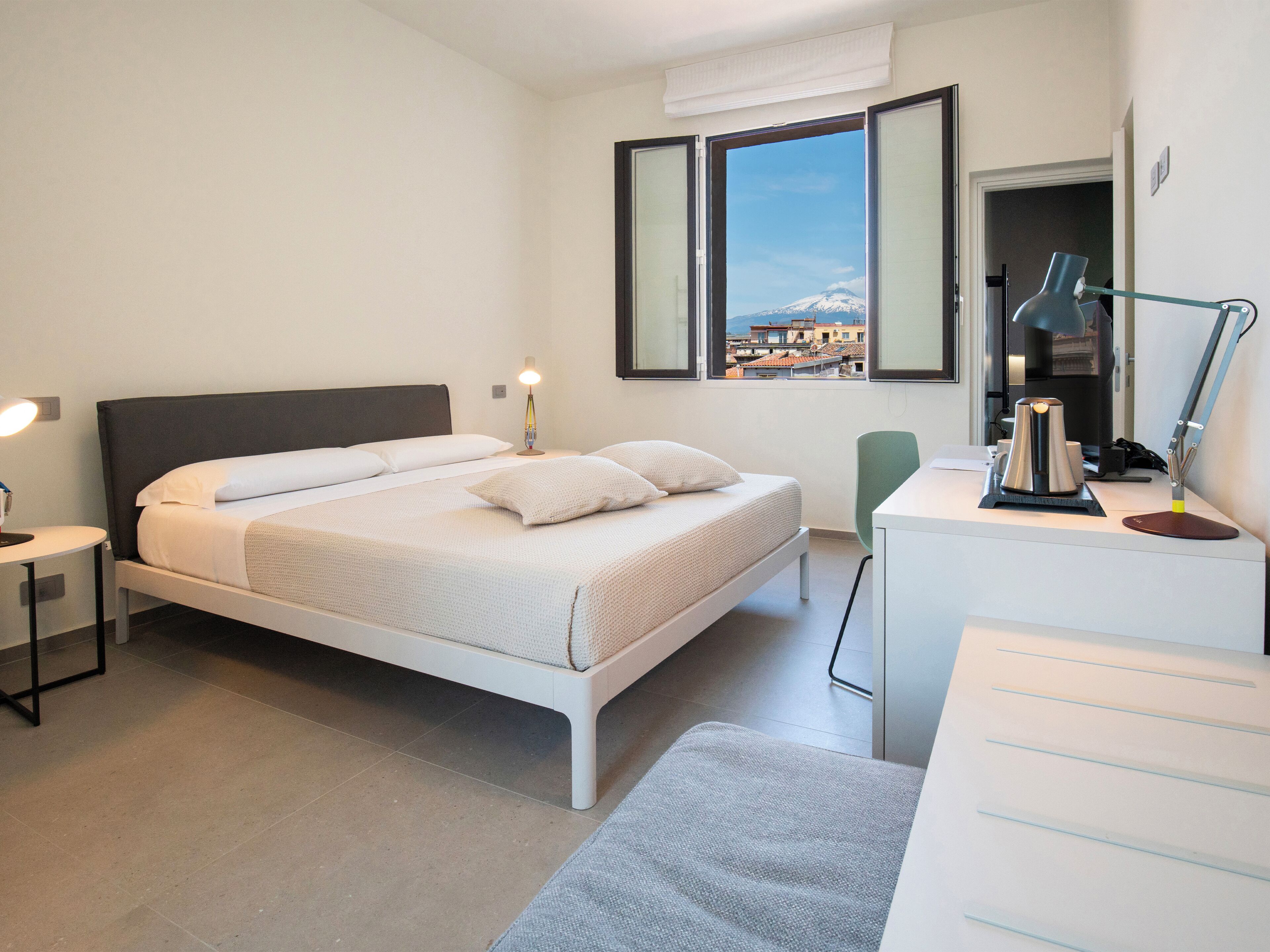 Un dormitorio moderno con cama, mesitas de noche, escritorio, silla y vista a una montaña a través de una ventana.