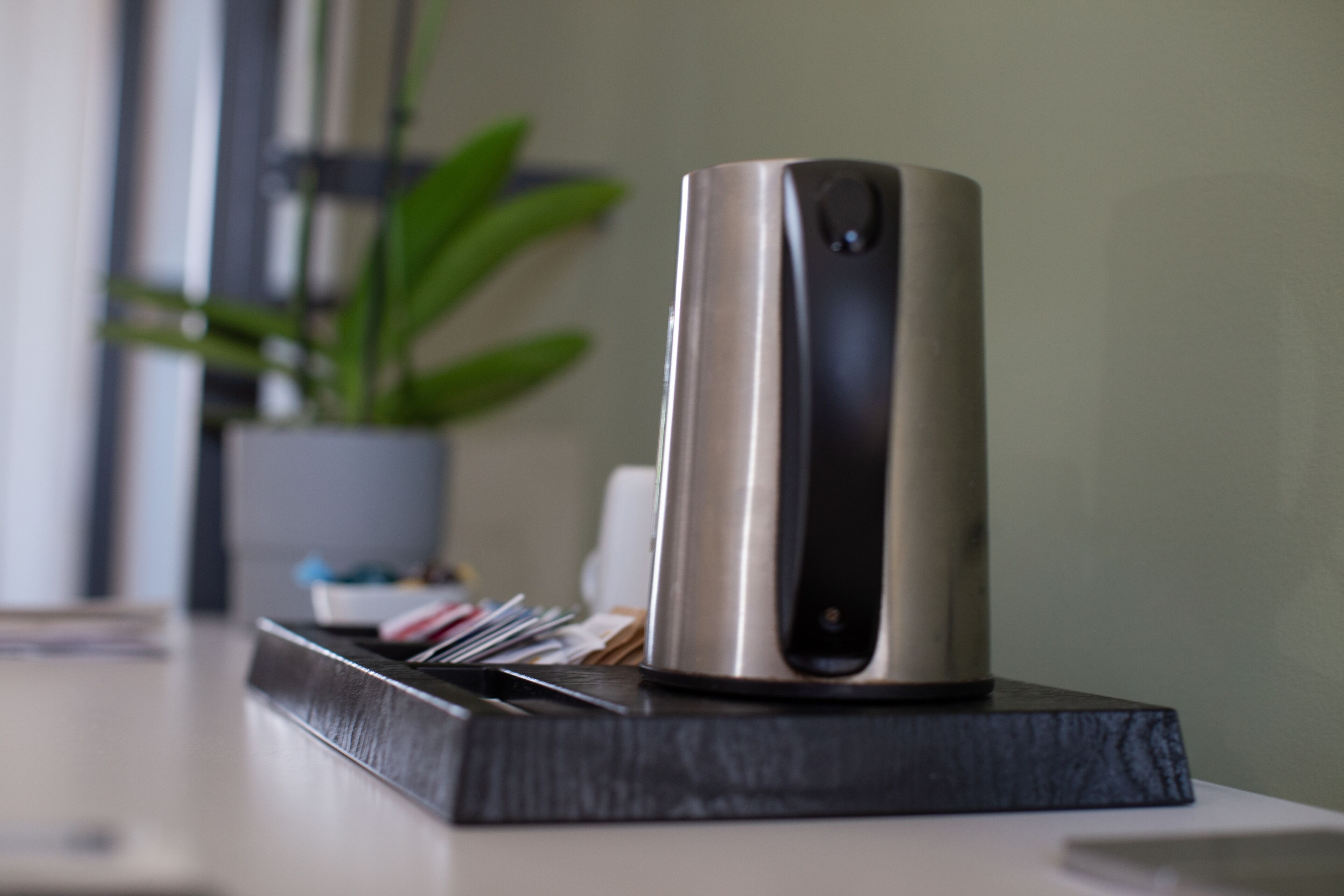 Een modern, metalen cilindervormig apparaat op een donker houten voet, waarschijnlijk een speaker of audioapparatuur, met een potplant op de achtergrond.