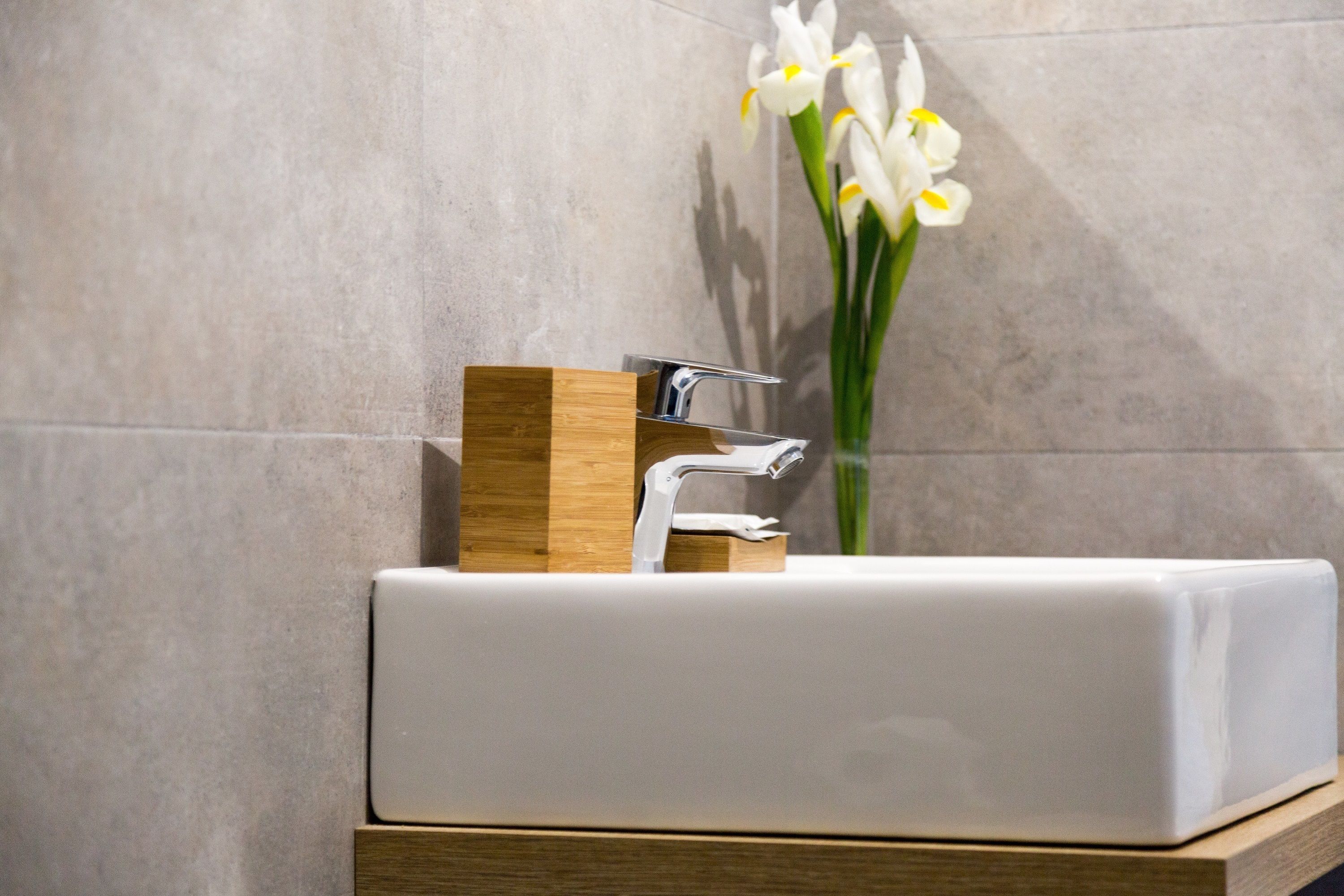 A modern bathroom with a white sink, wooden vanity, and a vase with white flowers.