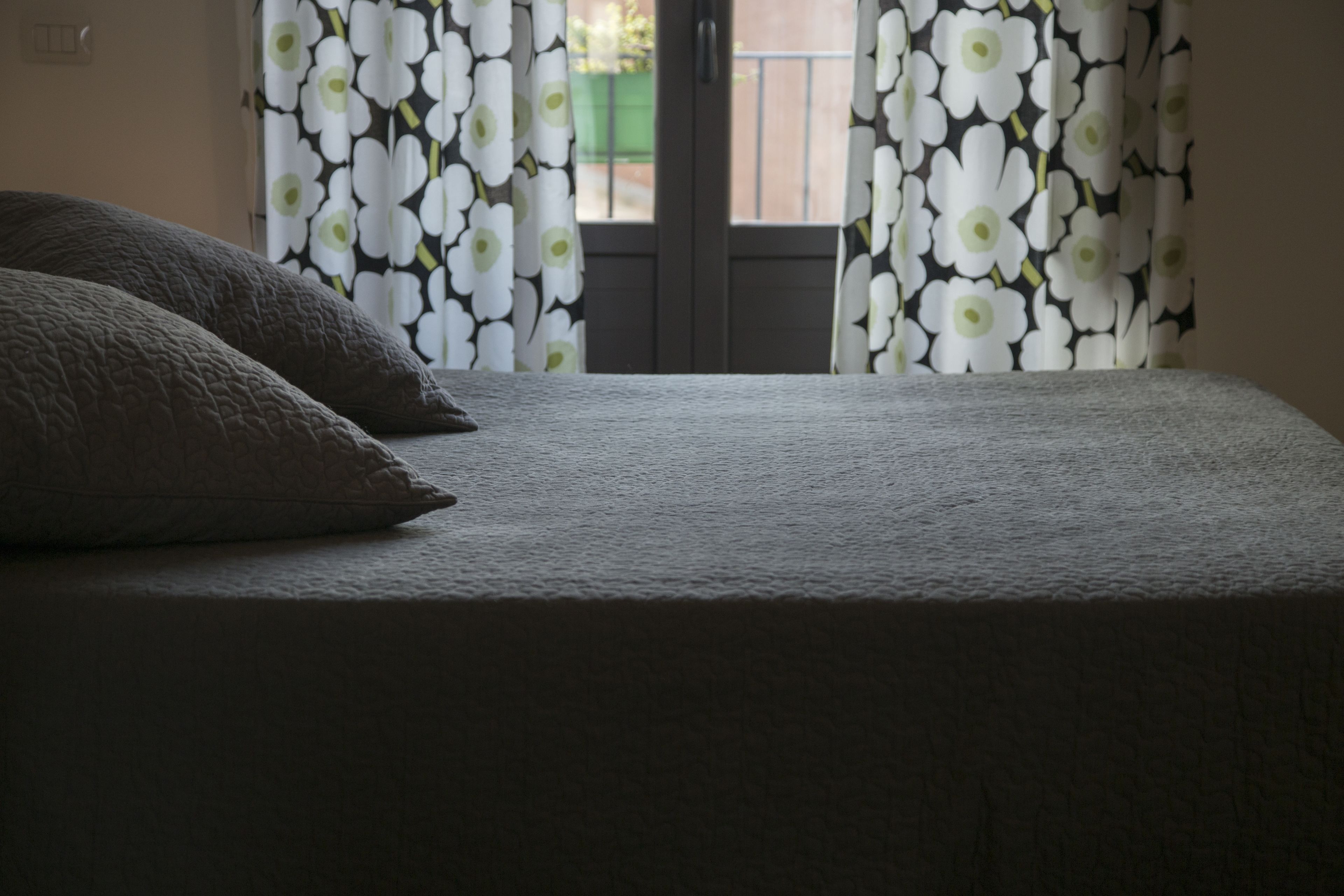 A bed with a textured blanket and two pillows, a window with floral curtains, and a view of a balcony outside.