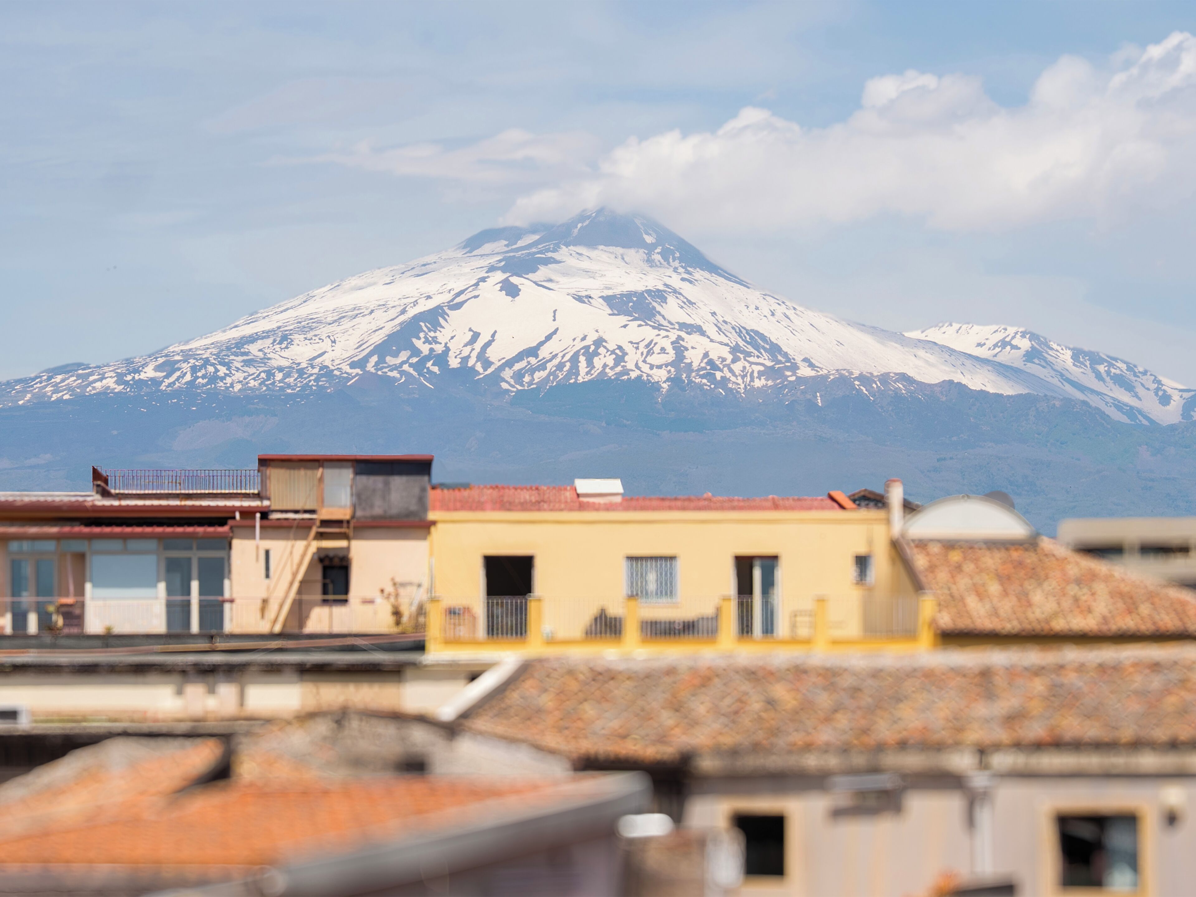 A mountain with snow-capped peaks in the background, a building with a red roof in the foreground, and another building with a yellow facade.