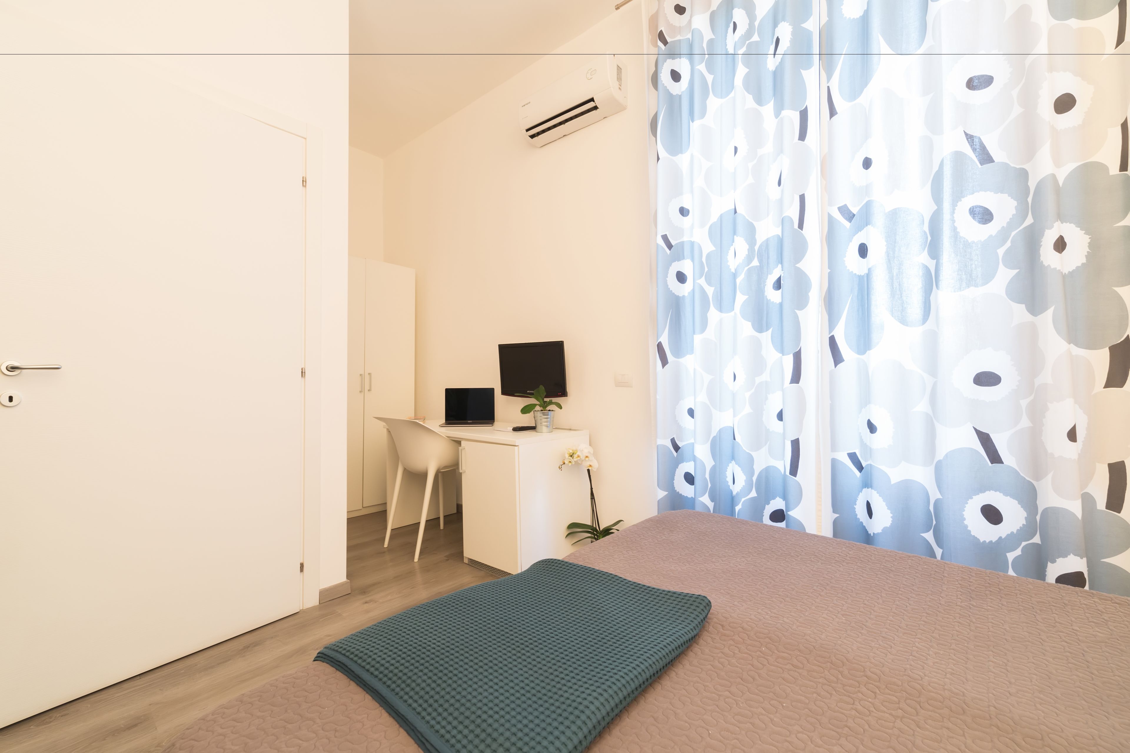 A bedroom with a bed, a desk with a computer, a chair, and a window with a floral curtain.