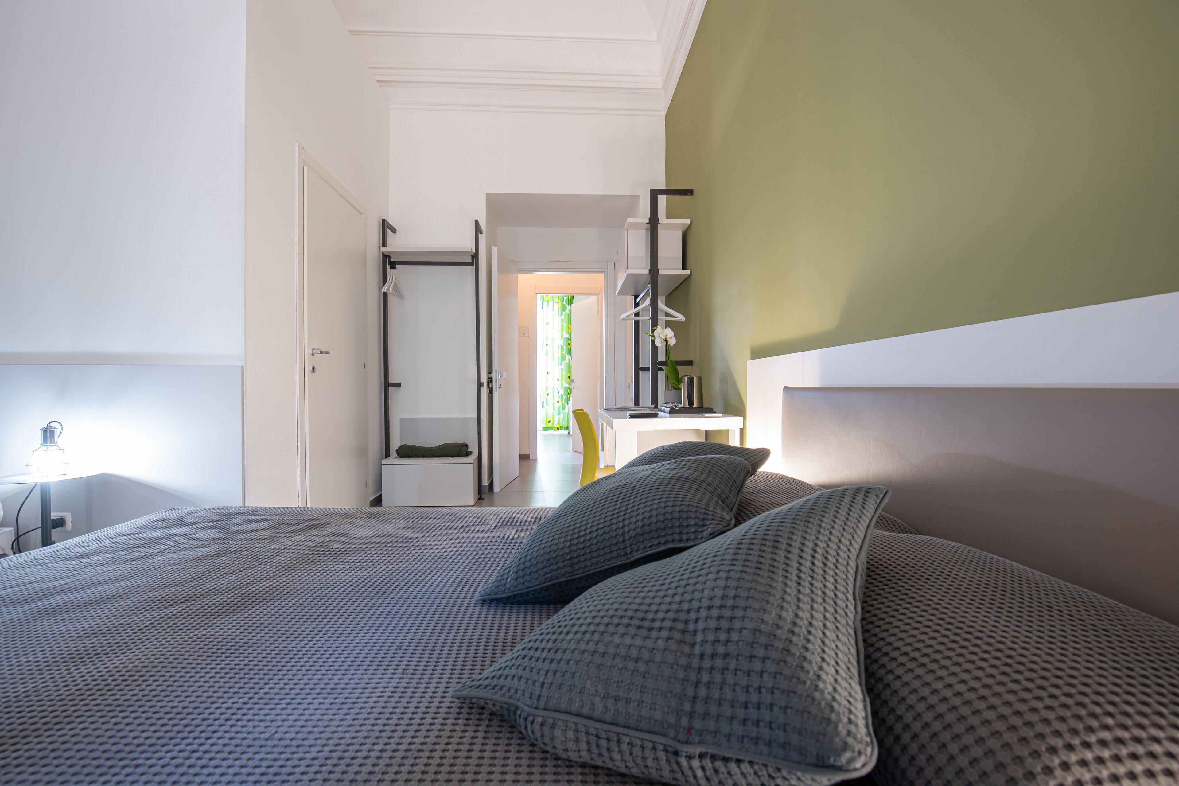 A neatly made bed with a patterned bedspread and pillows, a bedside lamp, and a view of a hallway leading to a room with a desk and chair.