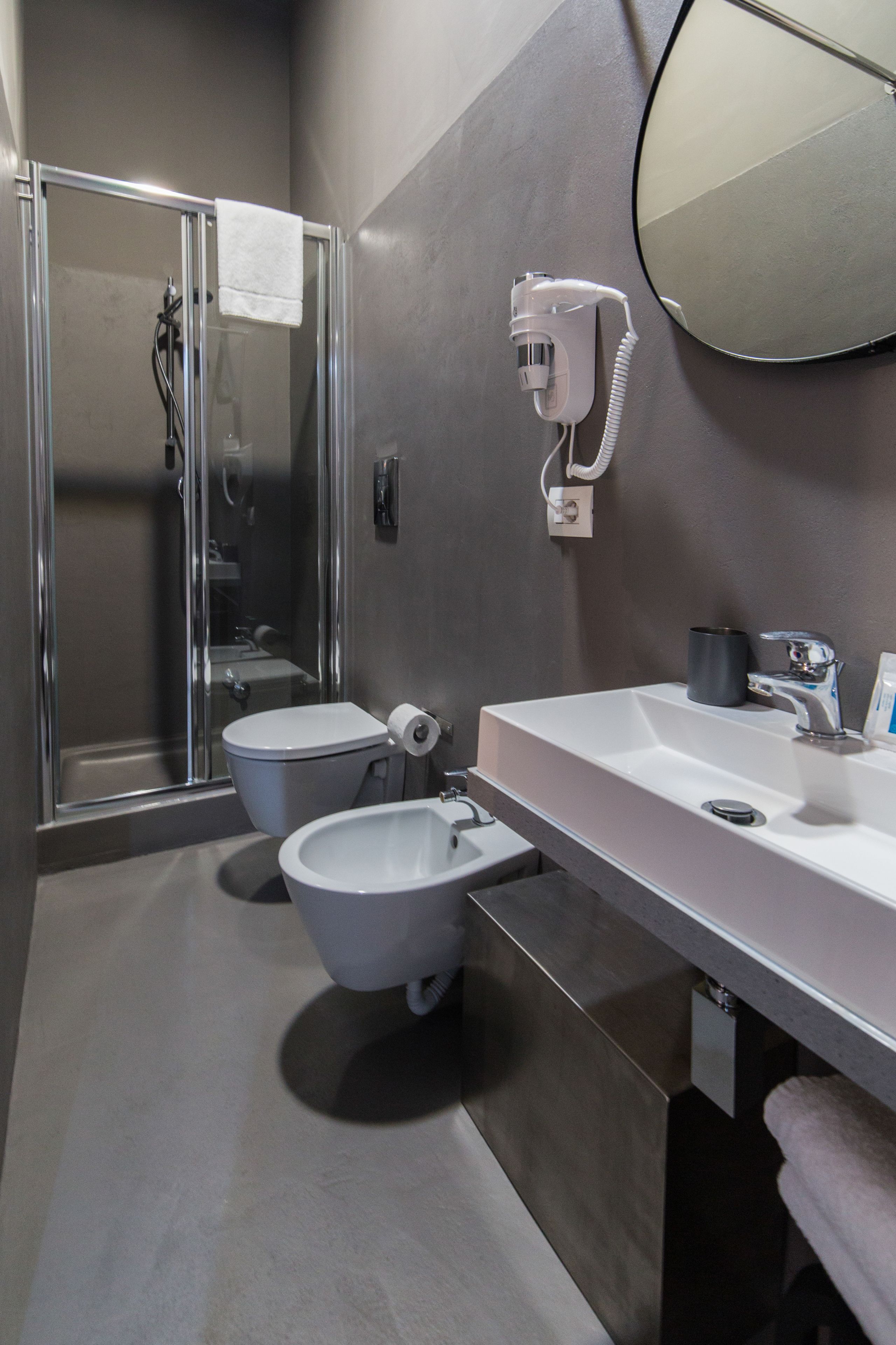 A modern bathroom with a glass shower enclosure, two toilets, a large mirror, and a hairdryer mounted on the wall.
