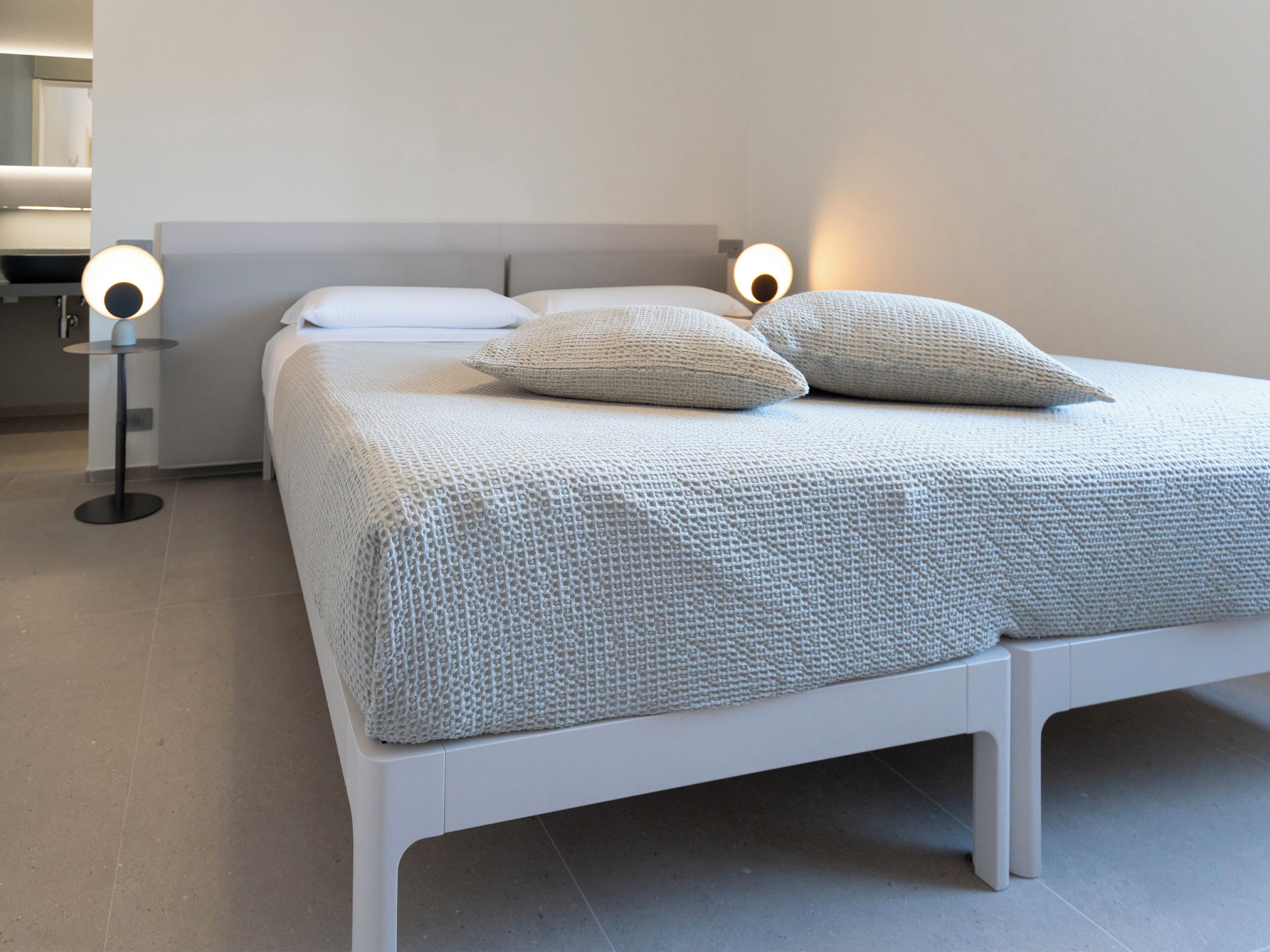 Una cama moderna con estructura blanca y una colcha texturizada, dos lámparas de noche y una mesita redonda.
