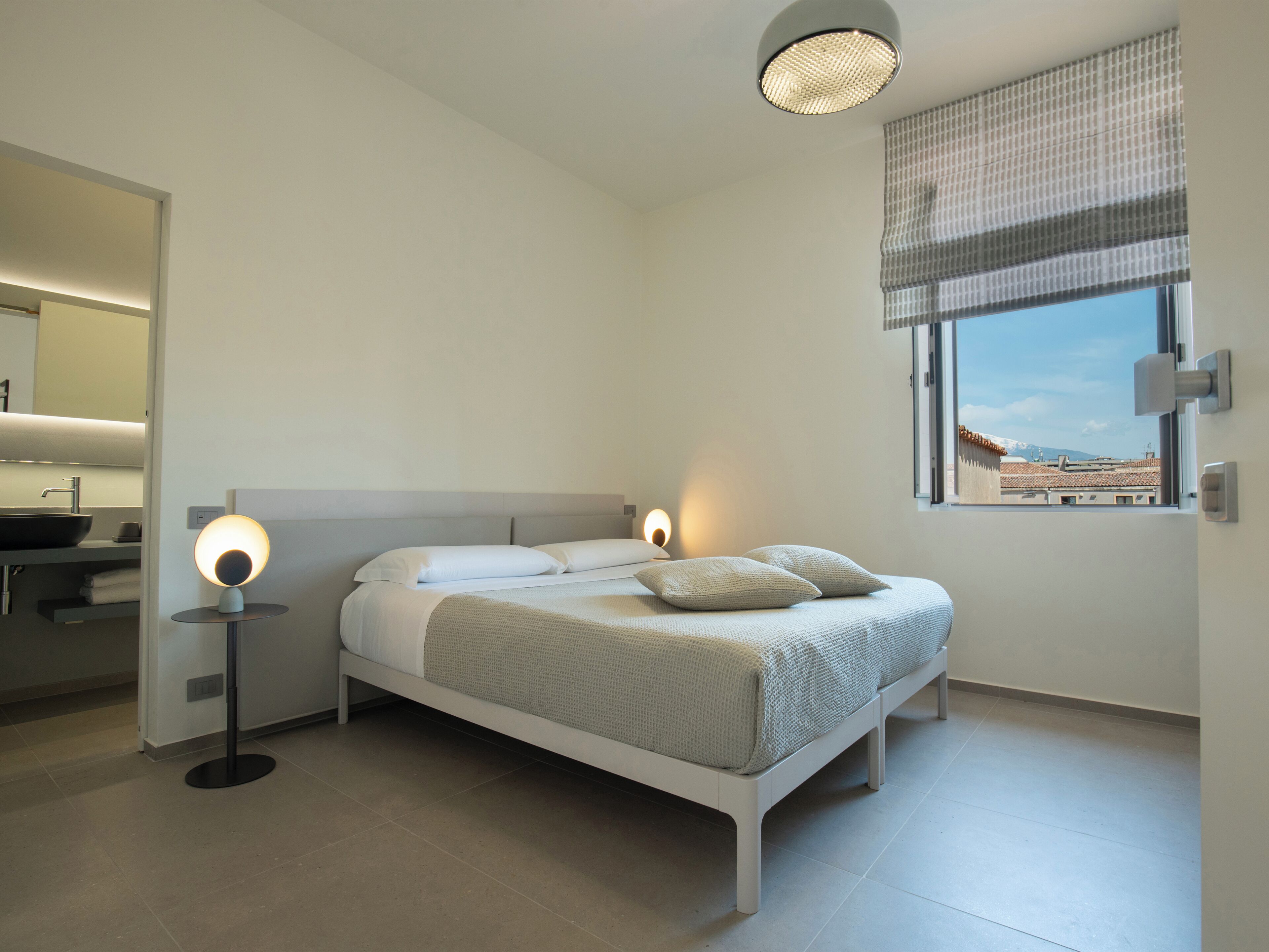 Camera da letto moderna con un letto, lampade da comodino, una finestra con vista e un bagno con lavandino e specchio.