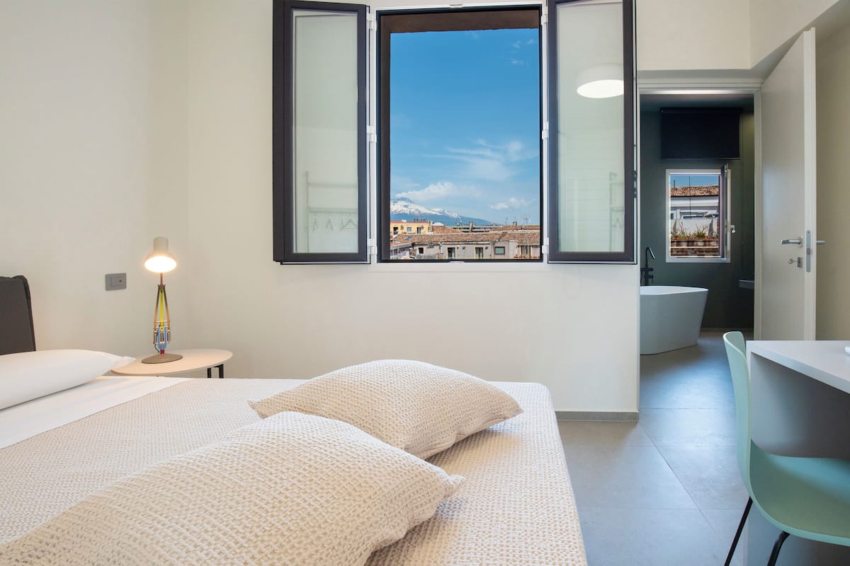 A modern hotel room with a large bed, a desk, a chair, a window with a city view, and a bathroom.