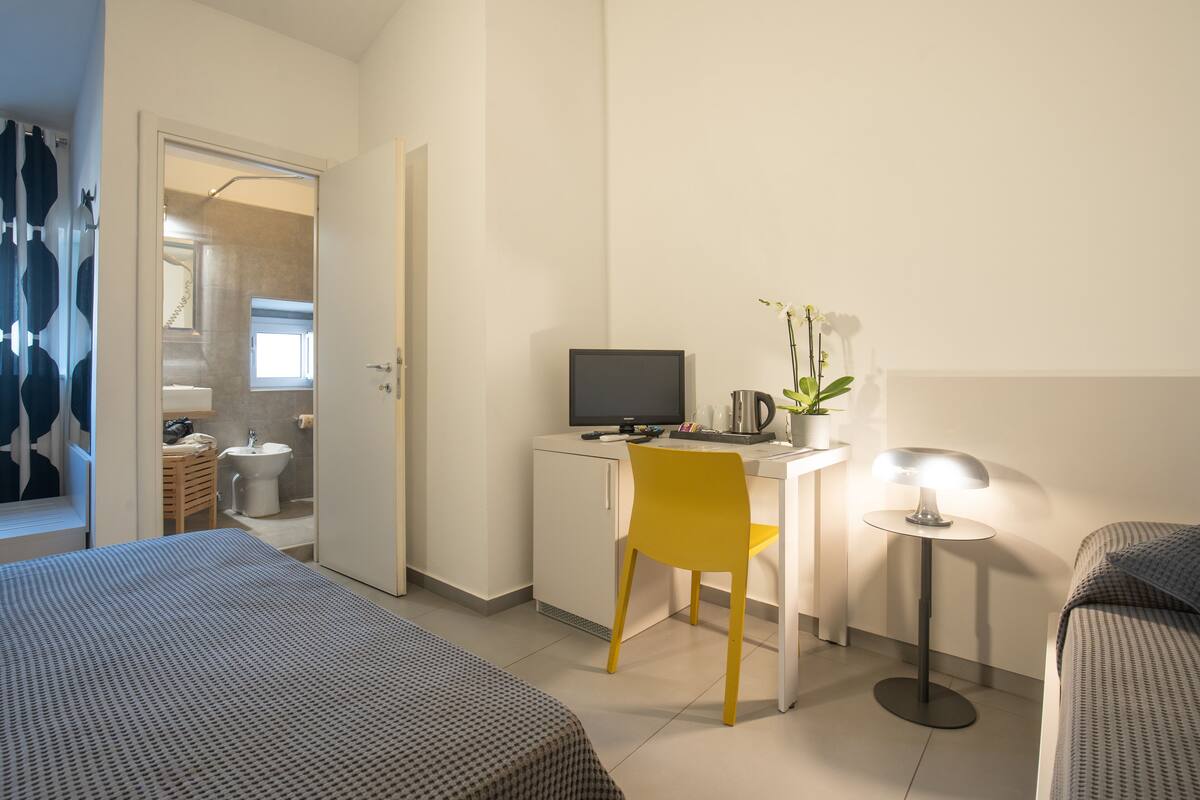 A hotel room with a bed, a desk with a computer, a chair, a lamp, and a small plant.